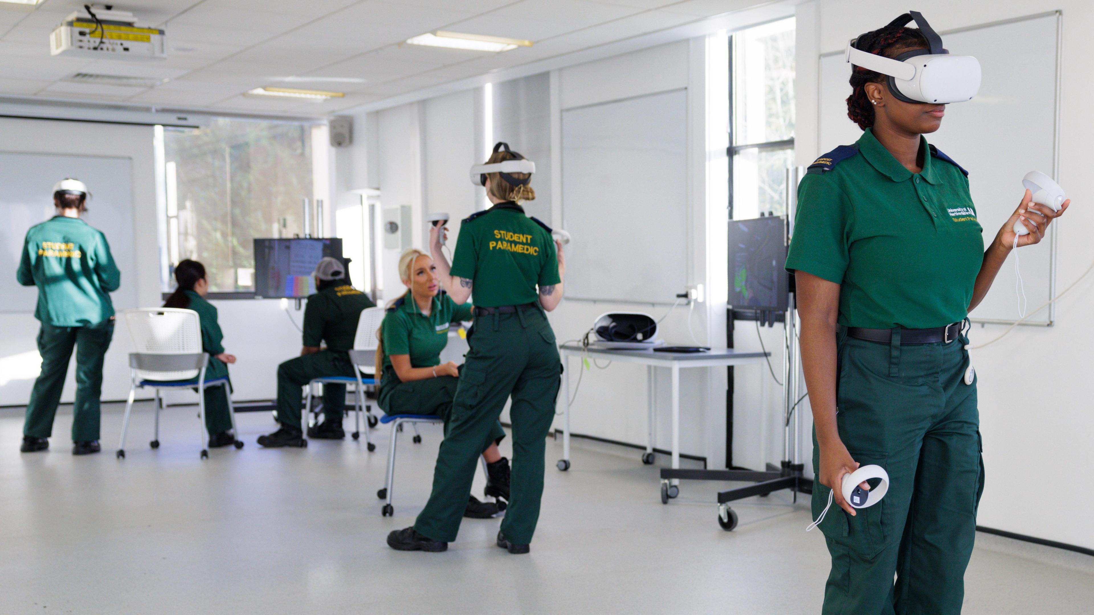 Six paramedic students taking part in a simulation session at university. They are all wearing a green uniform, with a top and trousers, with four wearing simulation headgear. They are in a while room, with monitors, chairs and desks with screens on them. 