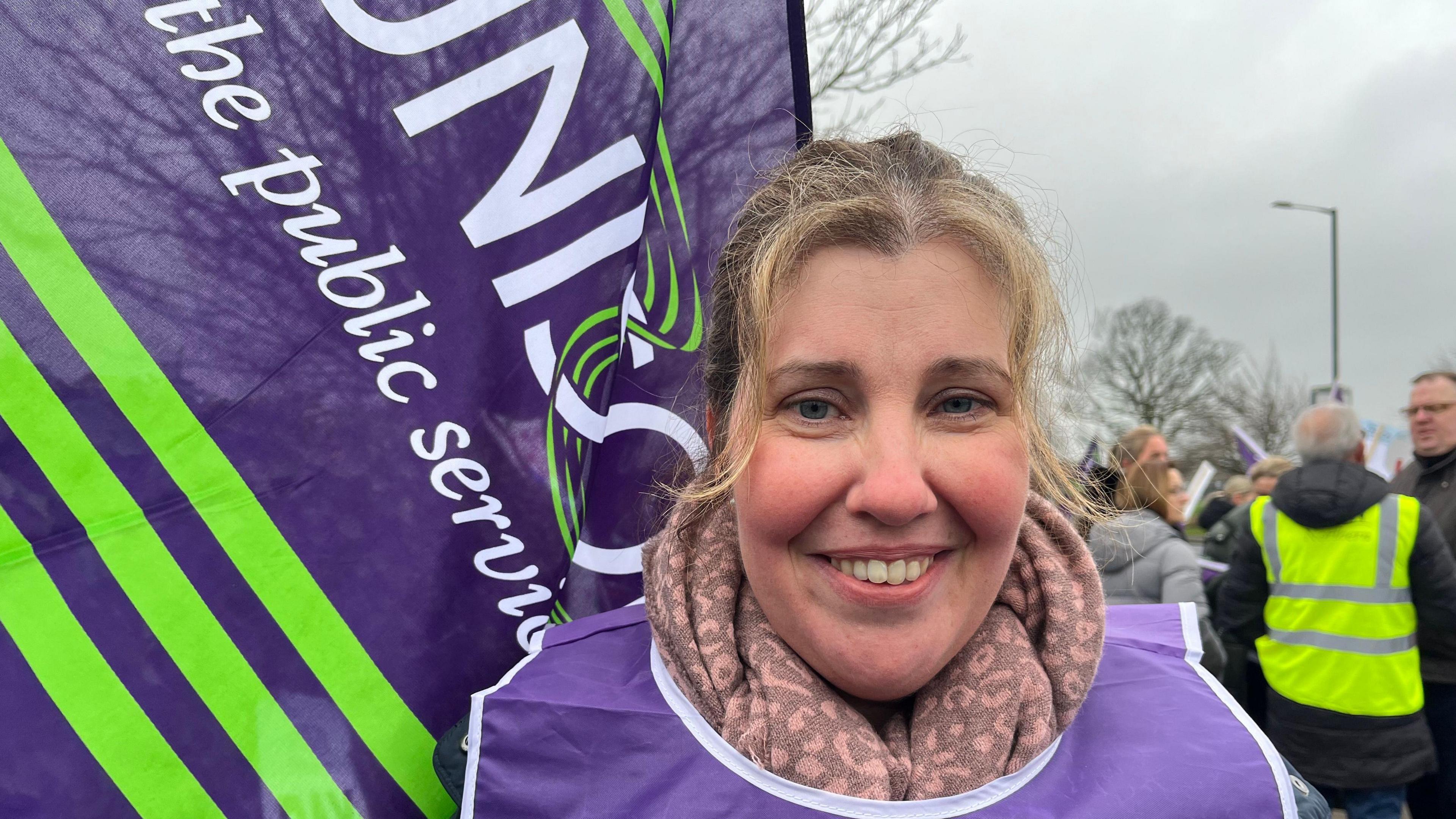 A healthcare worker on the picket line with a Unison flag behind her 