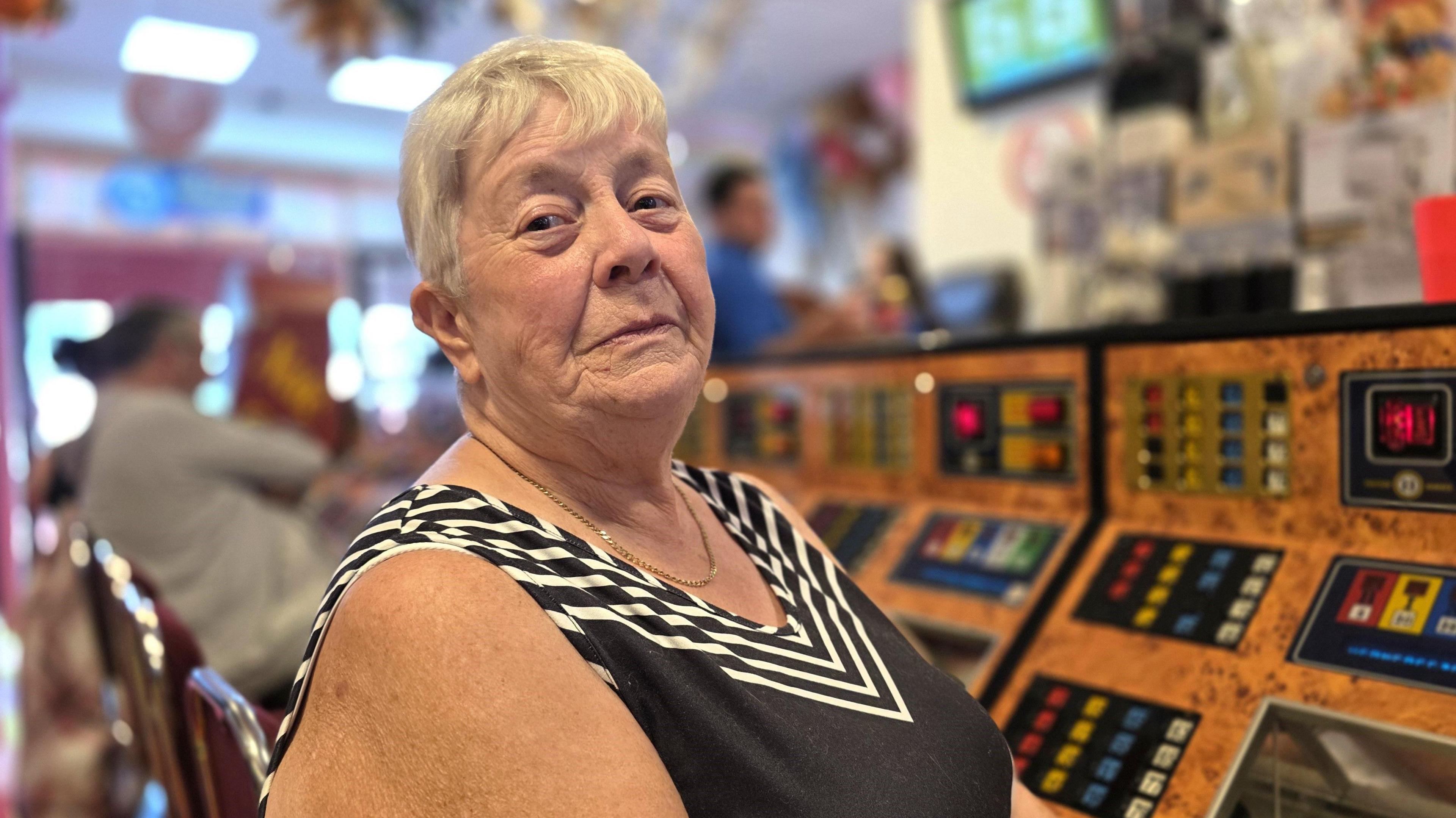Rita Fisher is sitting down - we can see she's at a bingo console. She's wearing a mostly black, sleeveless top - which has a white triangular pattern at the top.