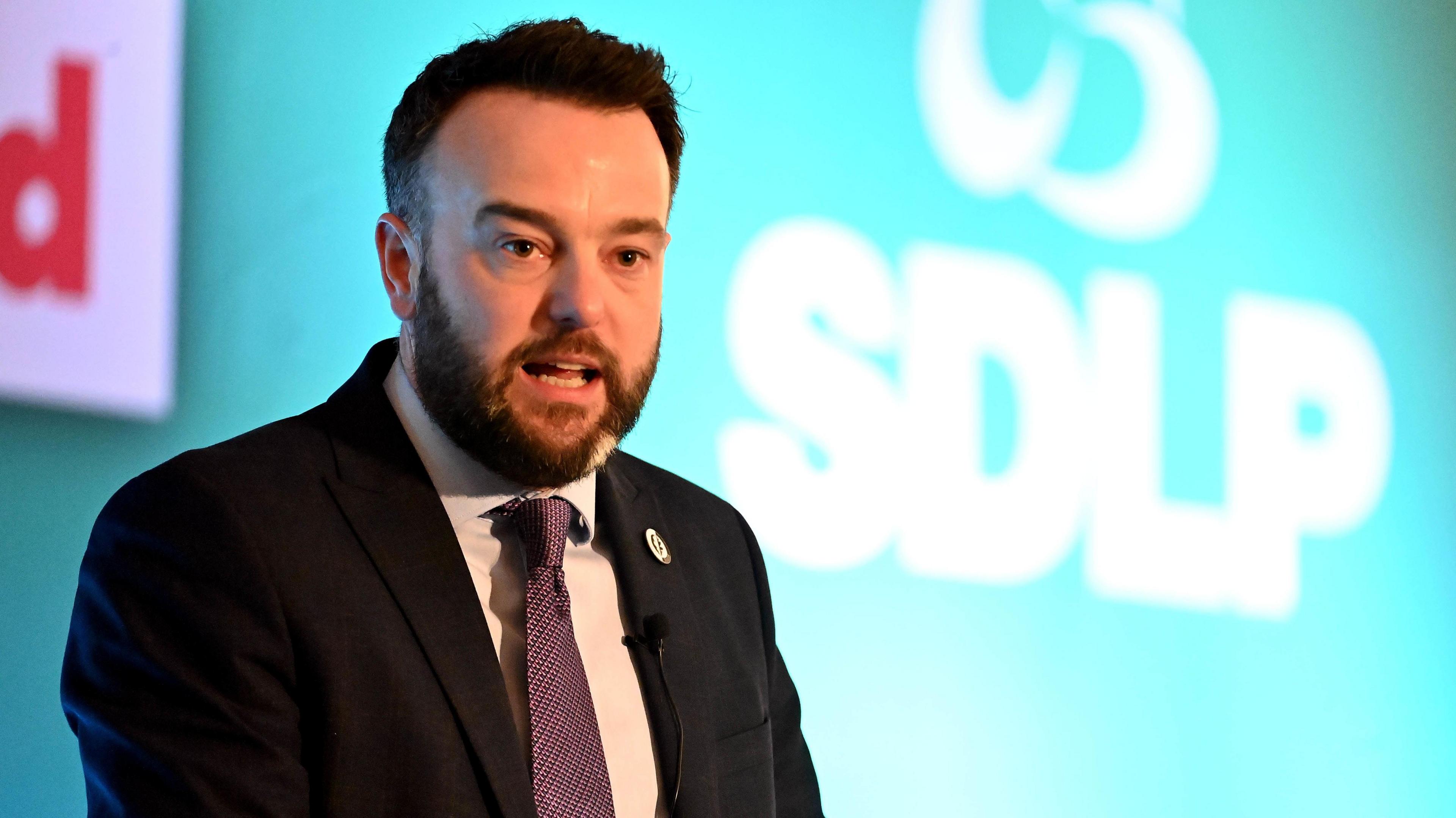 Colum Eastwood speaking in front of a backdrop that has SDLP branding. He is wearing a suit and tie