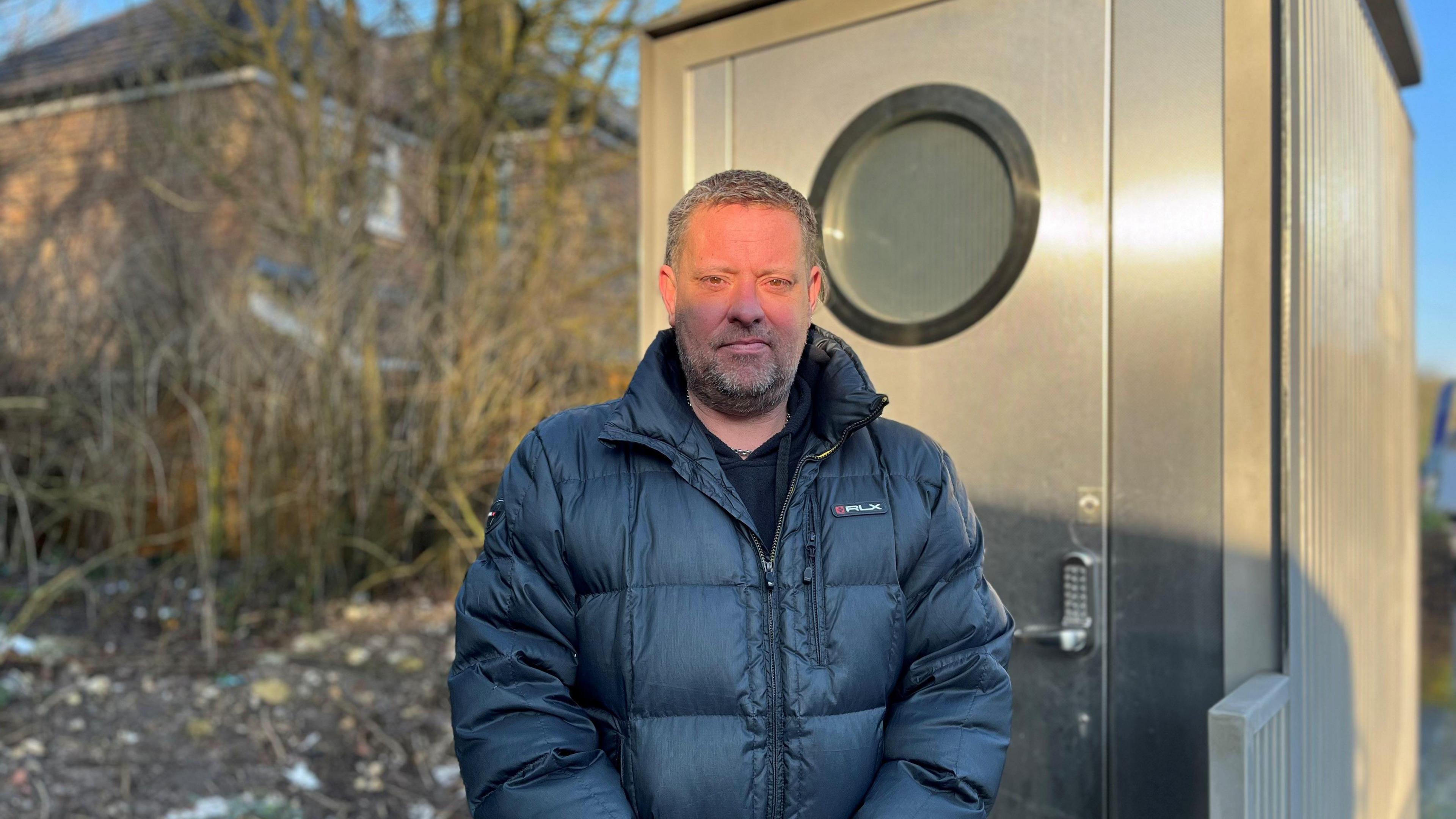 Brett Kemp, a man with short grey hair wearing a dark puffer jacket, standing outside a metal shed-style structure. It features a porthole window, corrugated sides, and a keypad-operated door handle. His house can be seen in the background behind a thin row of trees