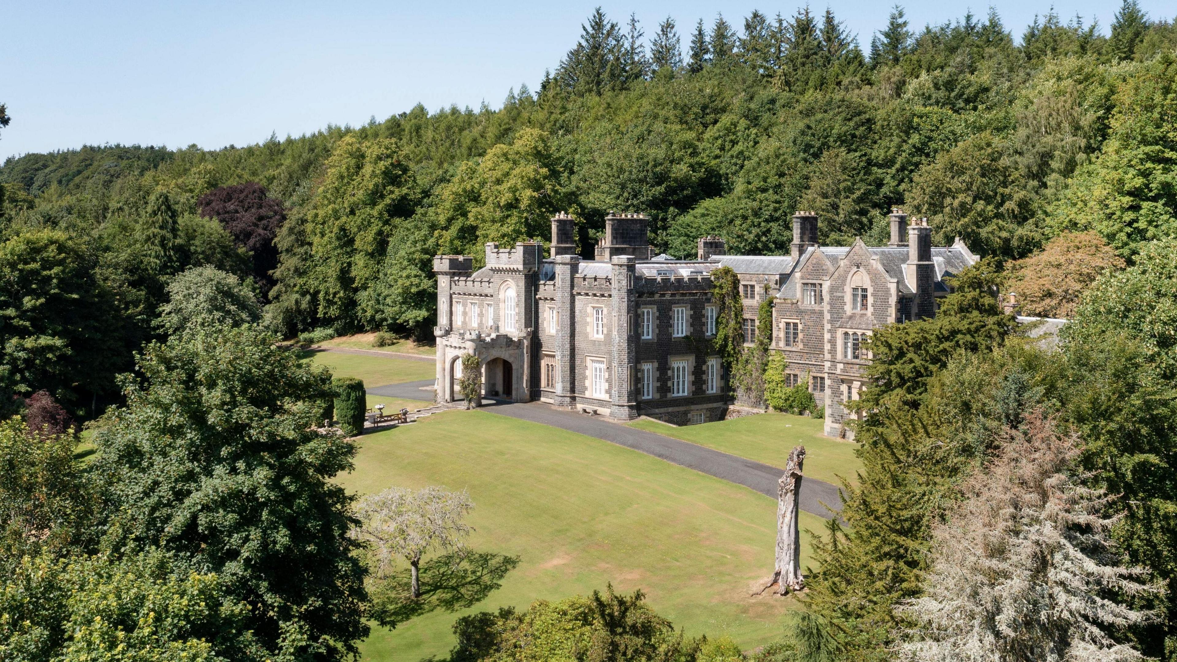 A grand stately home sits in a woodland opening. Surrounded mainly by tall green pine trees.