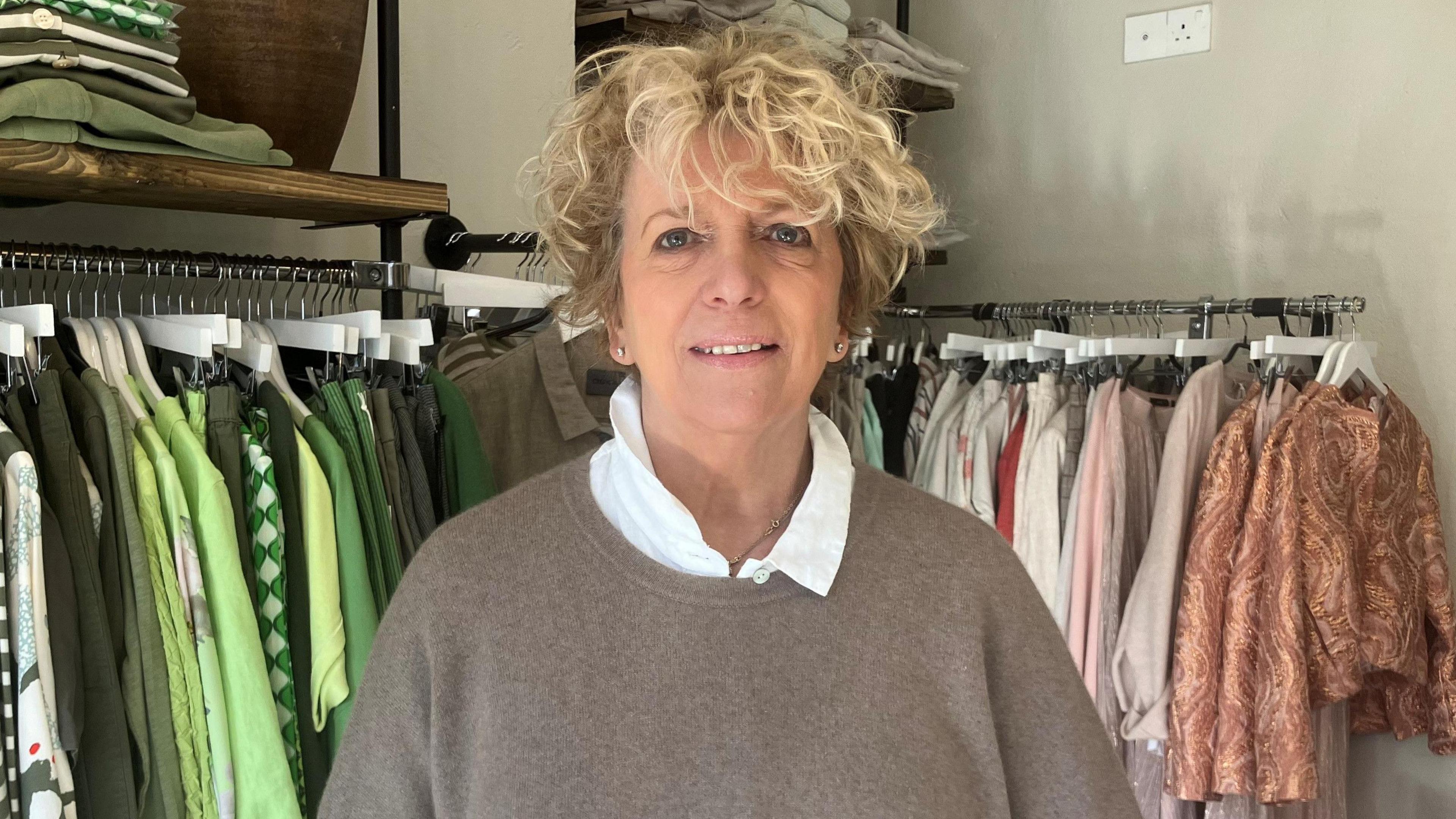 The owner of Nice Boutique in Rye, Lesley Farrow, stood in front of two rails of clothing stock. Lesley has dyed blonde hair and is wearing a grey jumper and white undershirt. 