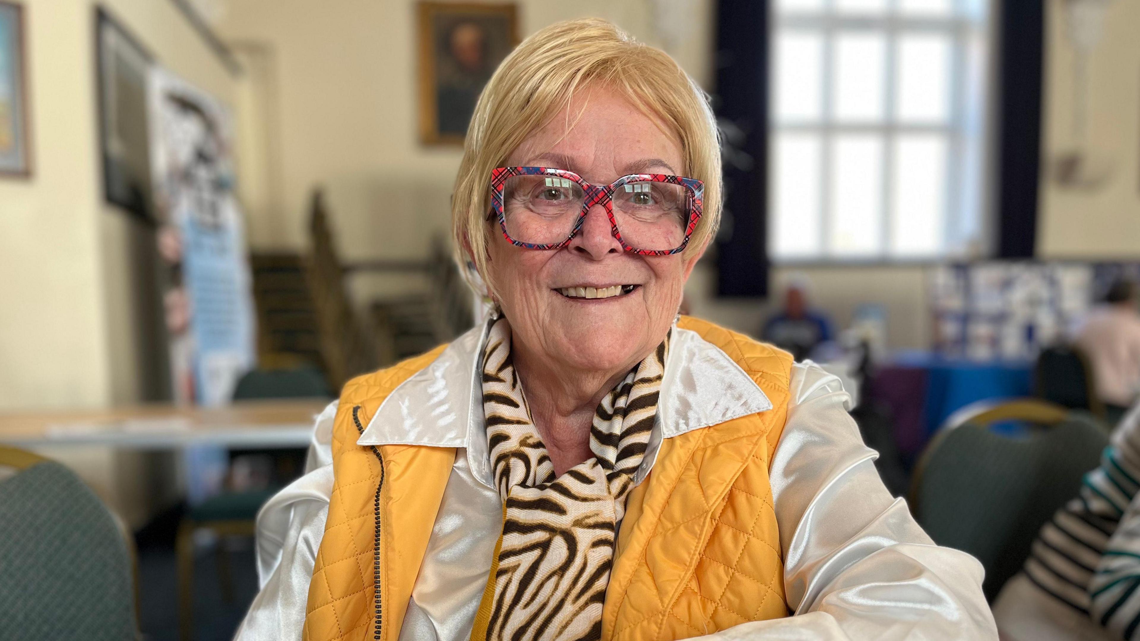 Hillary Letch has a blonde bob and red glasses. She is wearing a white shirt and yellow gilet.