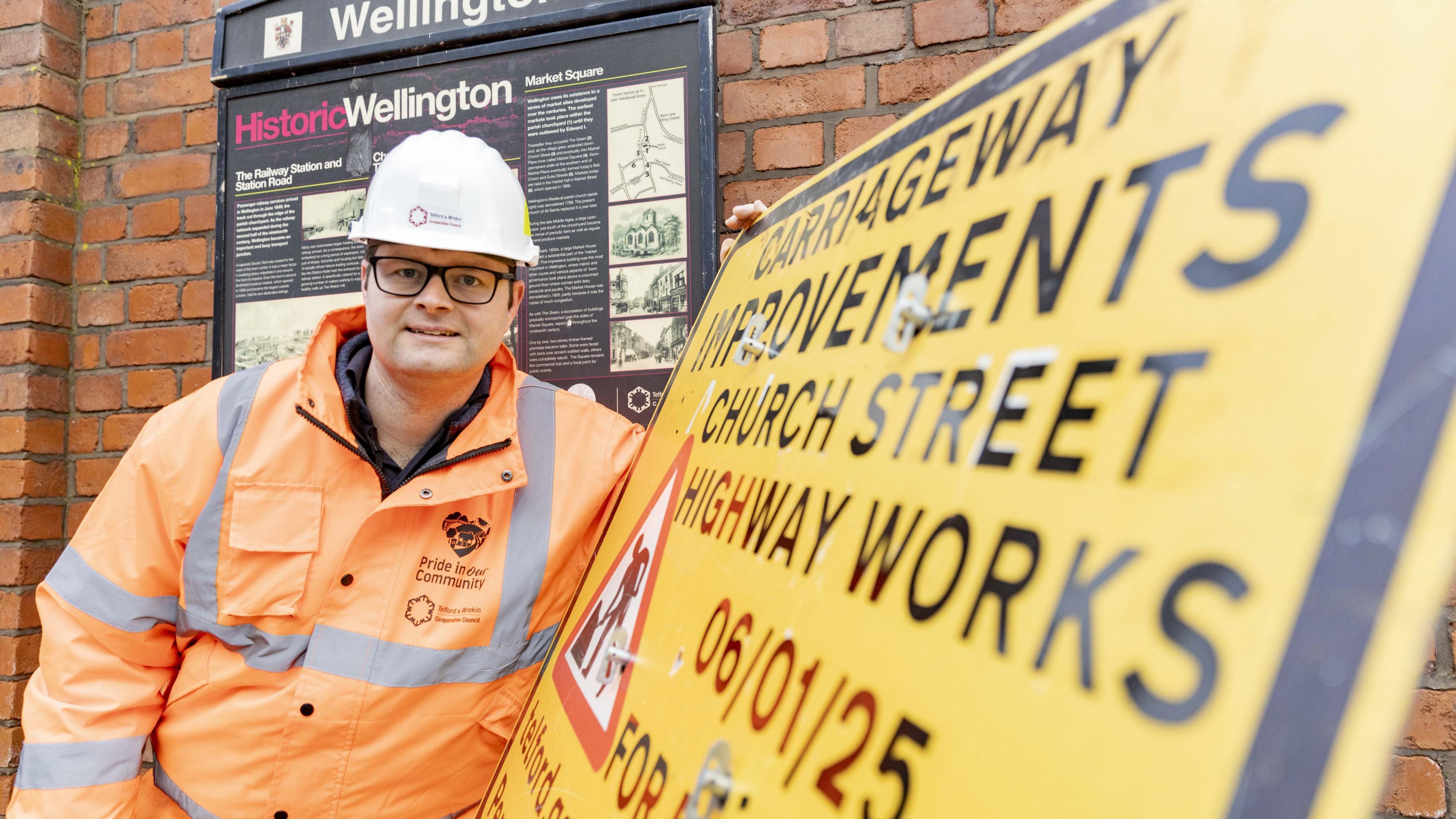 A man in a white hard hat and orange high-vis coat is leaning against a yellow roadworks sign. The sign says "carriageway improvements, church street highway works".