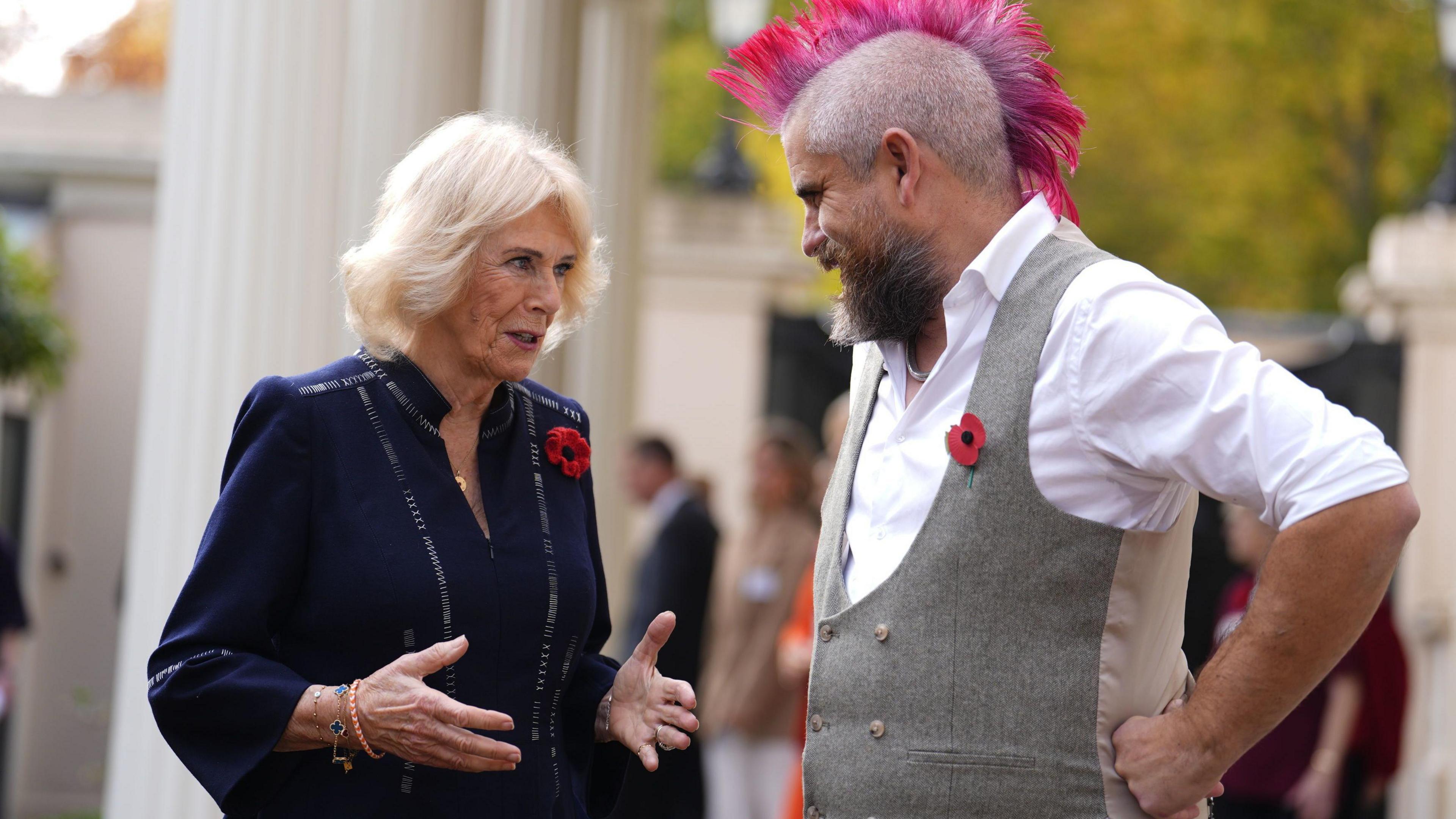 Queen Camilla, wearing a navy blue dress and a poppy, is seen talking to Ben Newman who has pink mohawk hair and is wearing a white shirt and grey waistcoat. They are standing outdoors with other people visible in the background.