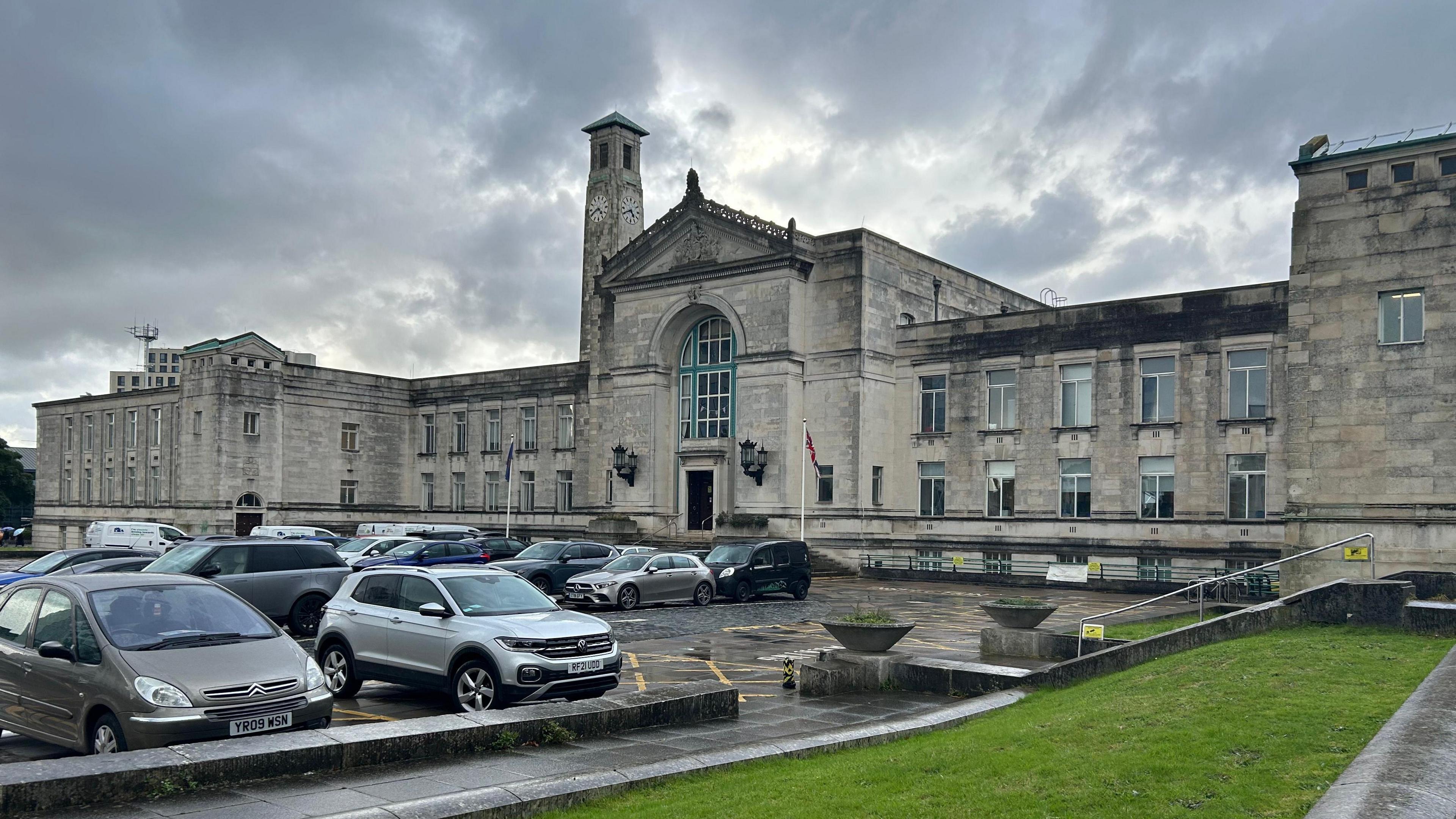 Southampton city council building. The large two-storey grey stone building has an overcast sky. There is a clock tower above the roof of the building and a car park outside with several vehicles parked in it. 