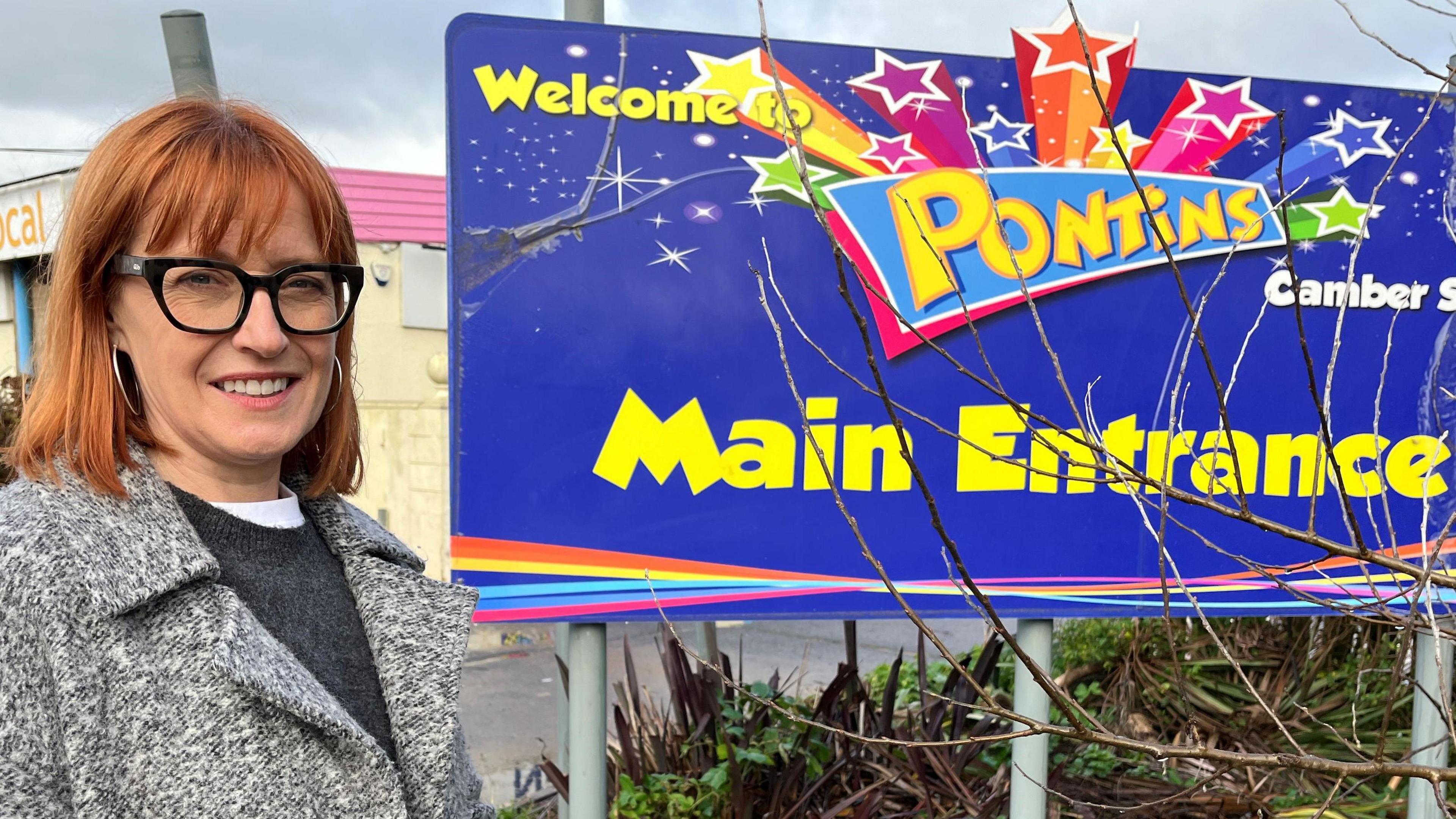 Sarah Broadbent is standing in from of the "Main Entrance" sign at the closed Pontin's holiday park. She is looking towards the camera. She has shoulder-length ginger hair with a fringe and black-rimmed glasses. She is wearing a dark grey jumper under a light grey coat.