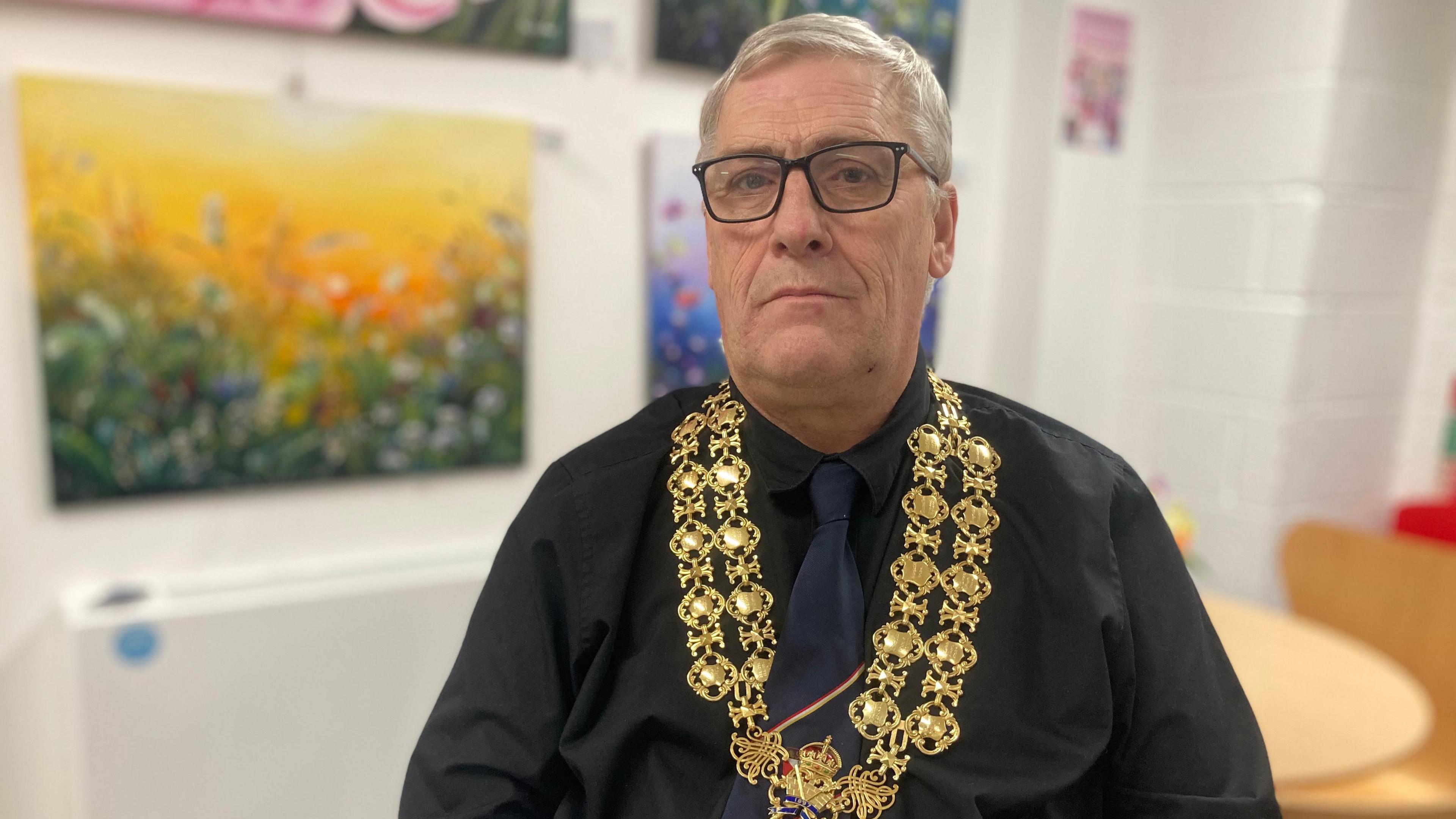 Councillor Phil Exton sitting down in a room with colourful landscape paintings behind him. He has short white hair and is wearing a black shirt, black tie, and black square framed glasses. Around his neck are his mayoral regalia, two gold chains with shields on them.