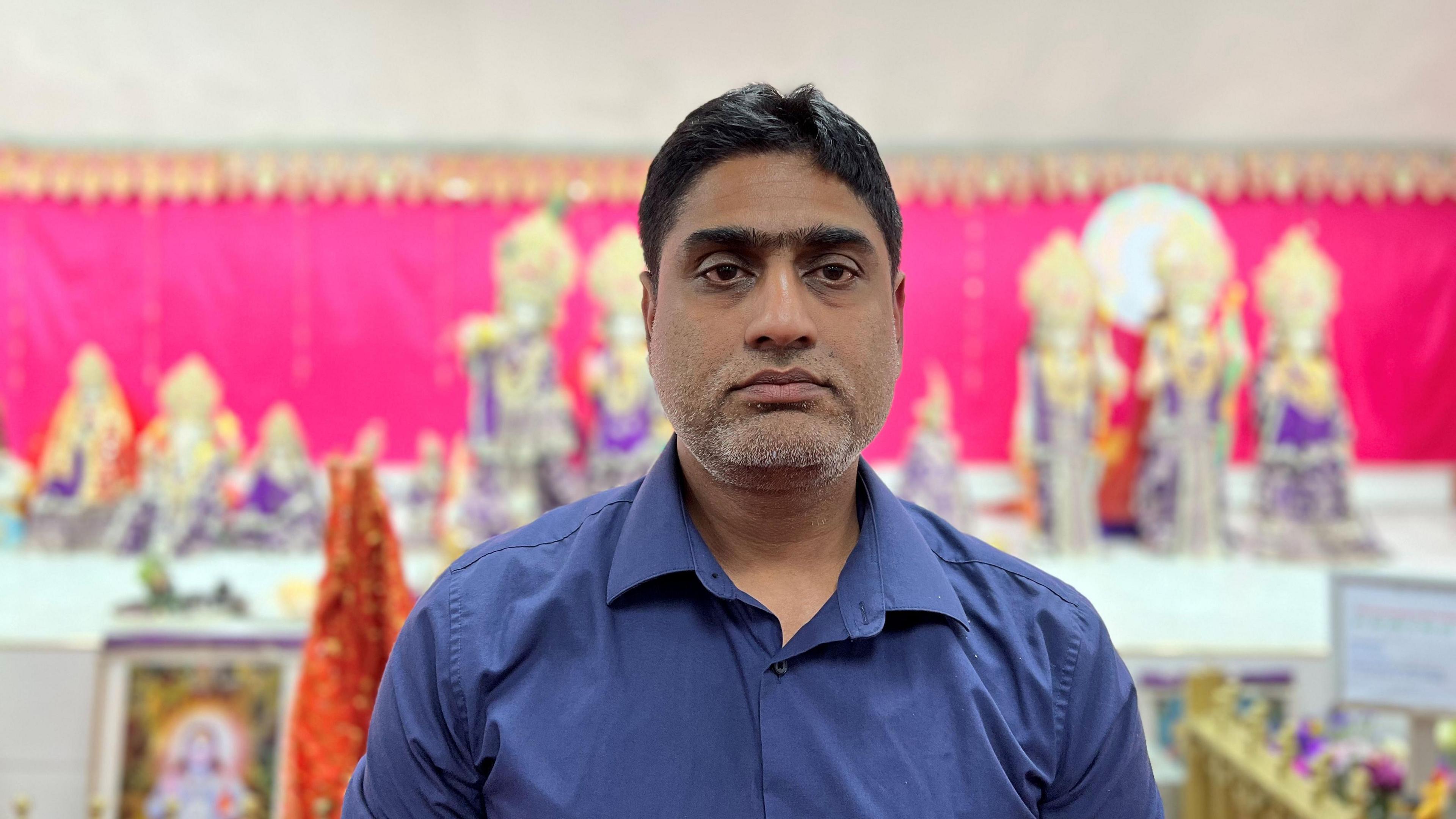 Dr Rajesh Gurrala has short black hair and a blue open-collared shirt, he is standing in front of a pink backdrop and colourful pictures and statues