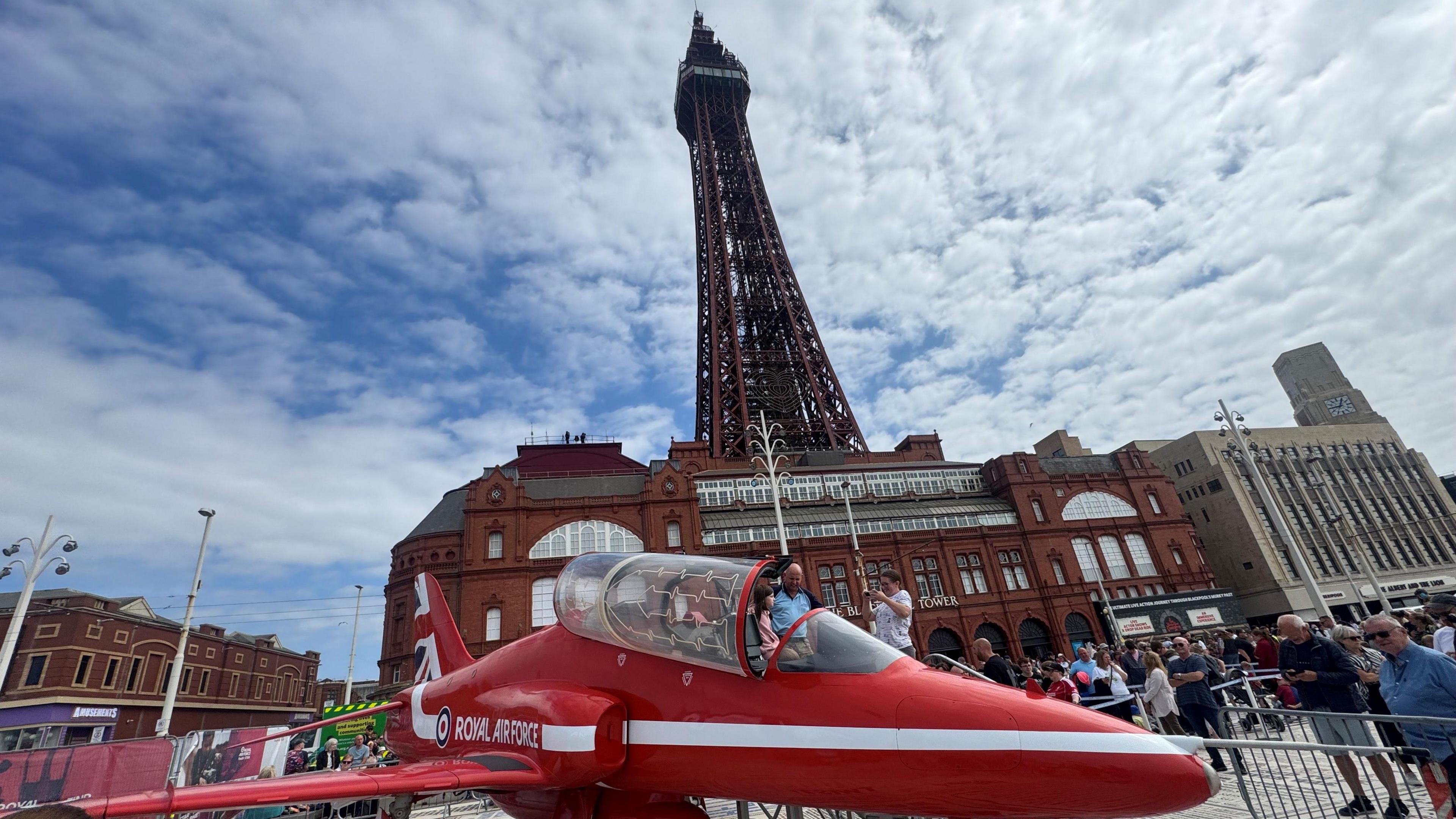 A red RAF plane with some visitors inside the cockpit is surrounded by a crowd of onlookers on the seafront. Blackpool Tower rises up in the background with white clouds in the sky above.