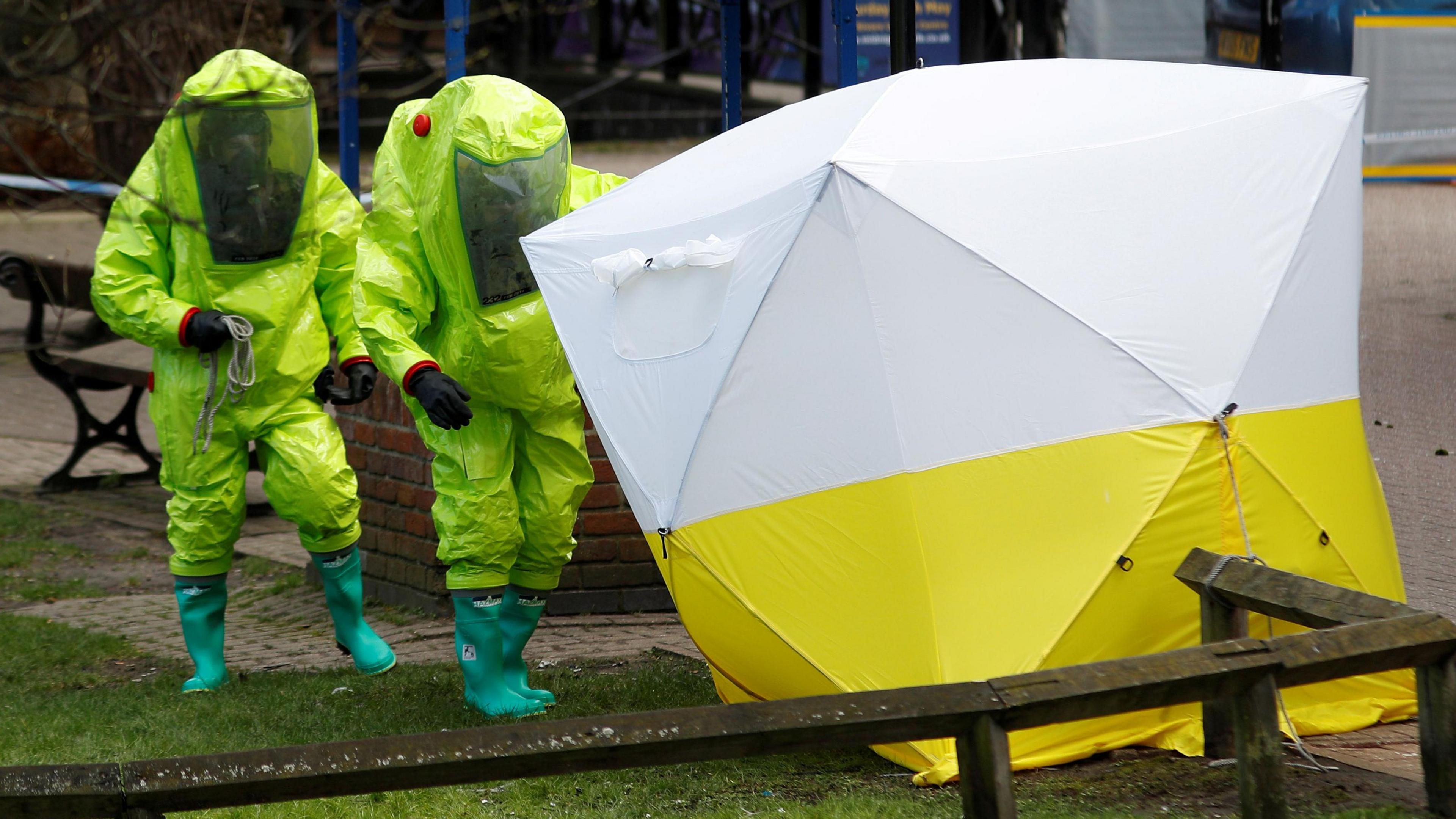 Two people in green hazmat suits next to a white and yellow forensic tent. 