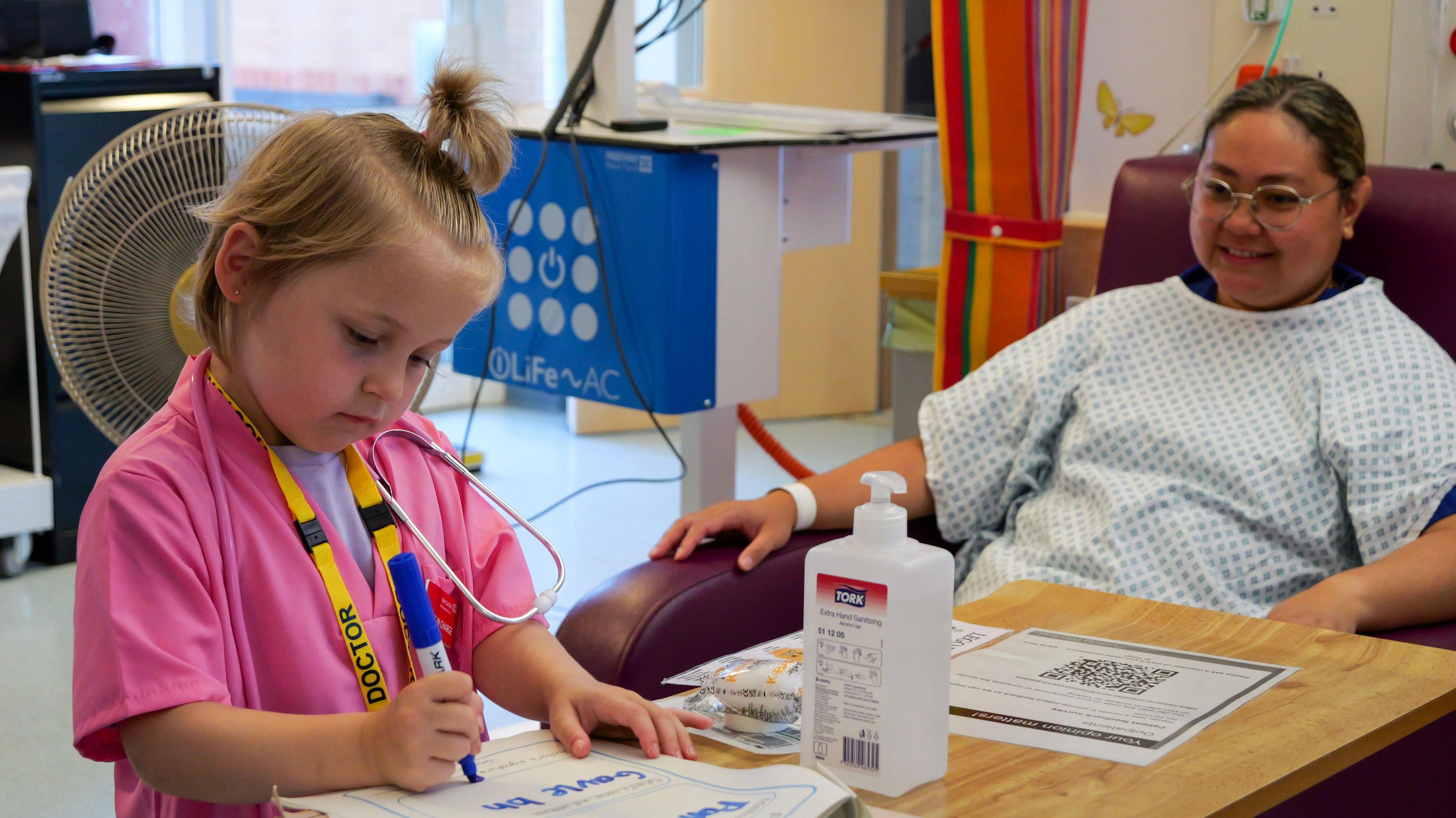 A young blonde girl in pink doctors' scrubs writing on a piece of paper that is on a wooden table that is positioned in front of a hospital patient. 