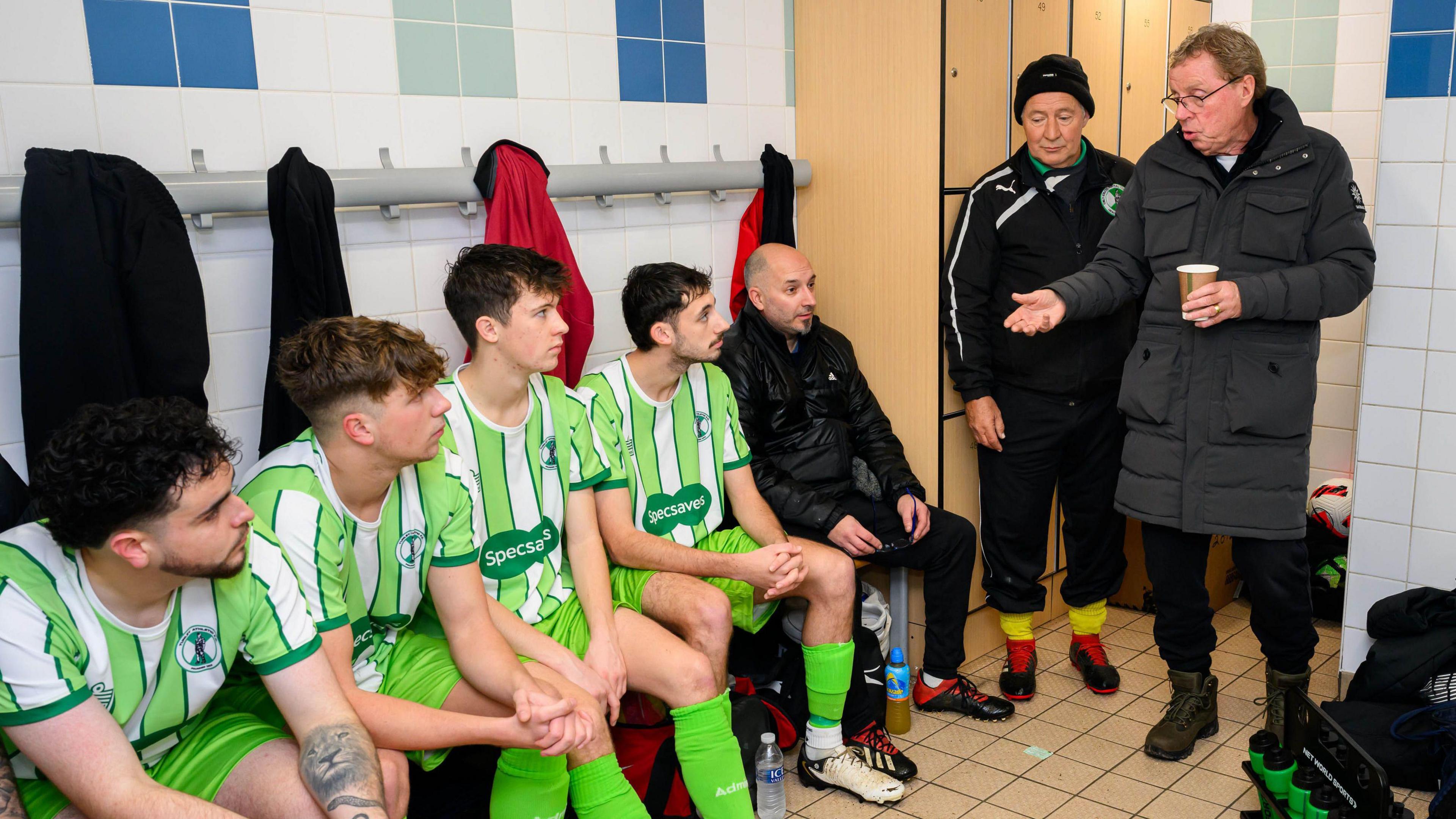 Harry Redknapp is standing on the right of the picture, pointing at players of Tunley Athletic FC inside the club's dressing room. The players are wearing green jerseys and are looking at Mr Redknapp.