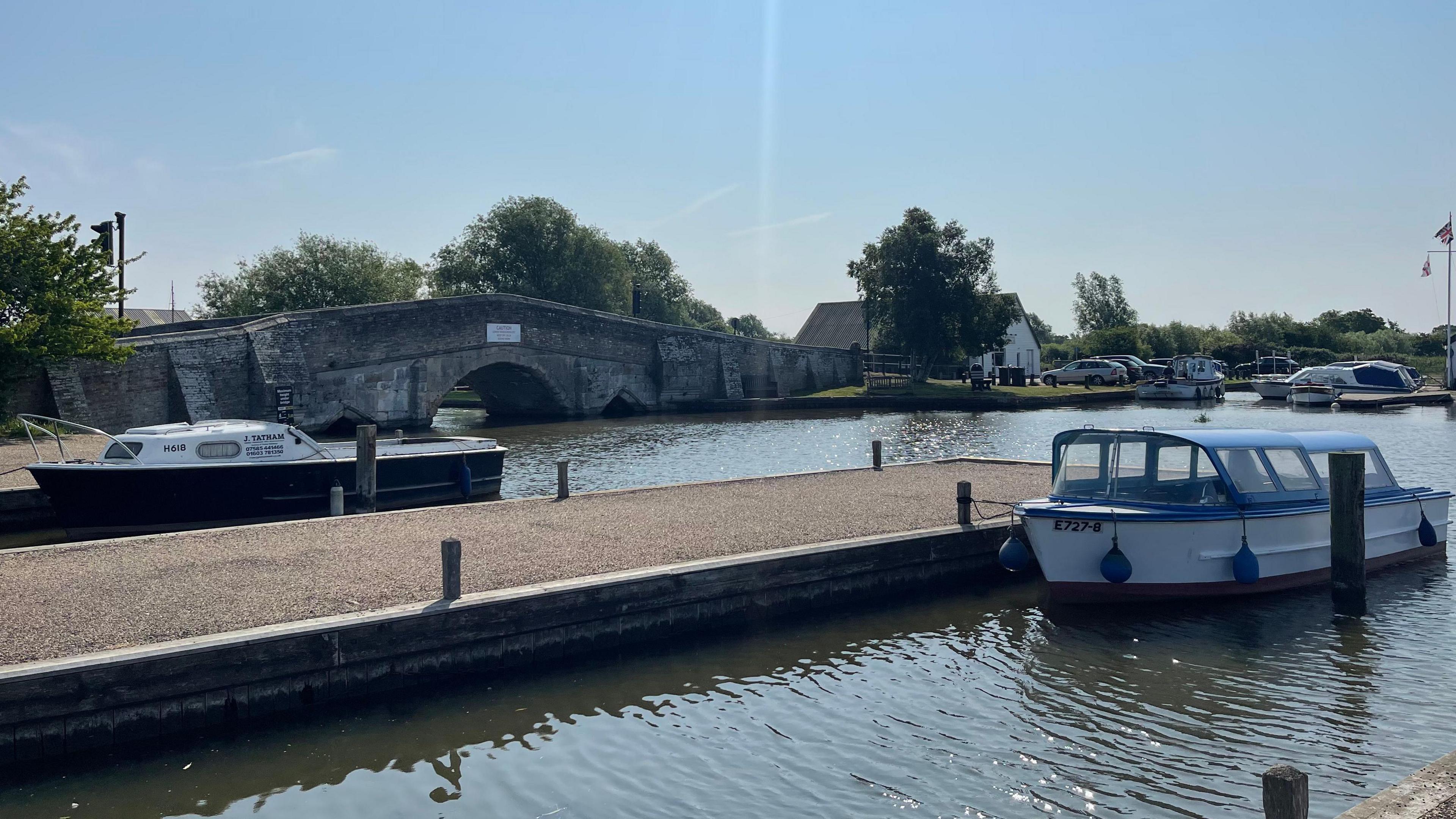 Two blue-and-white boats moored on the Broads. Behind them, a wide stretch of water goes under a bridge, which is on the left of the picture. In the distance, on the other side of the river, buildings , cars and boots can be seen.