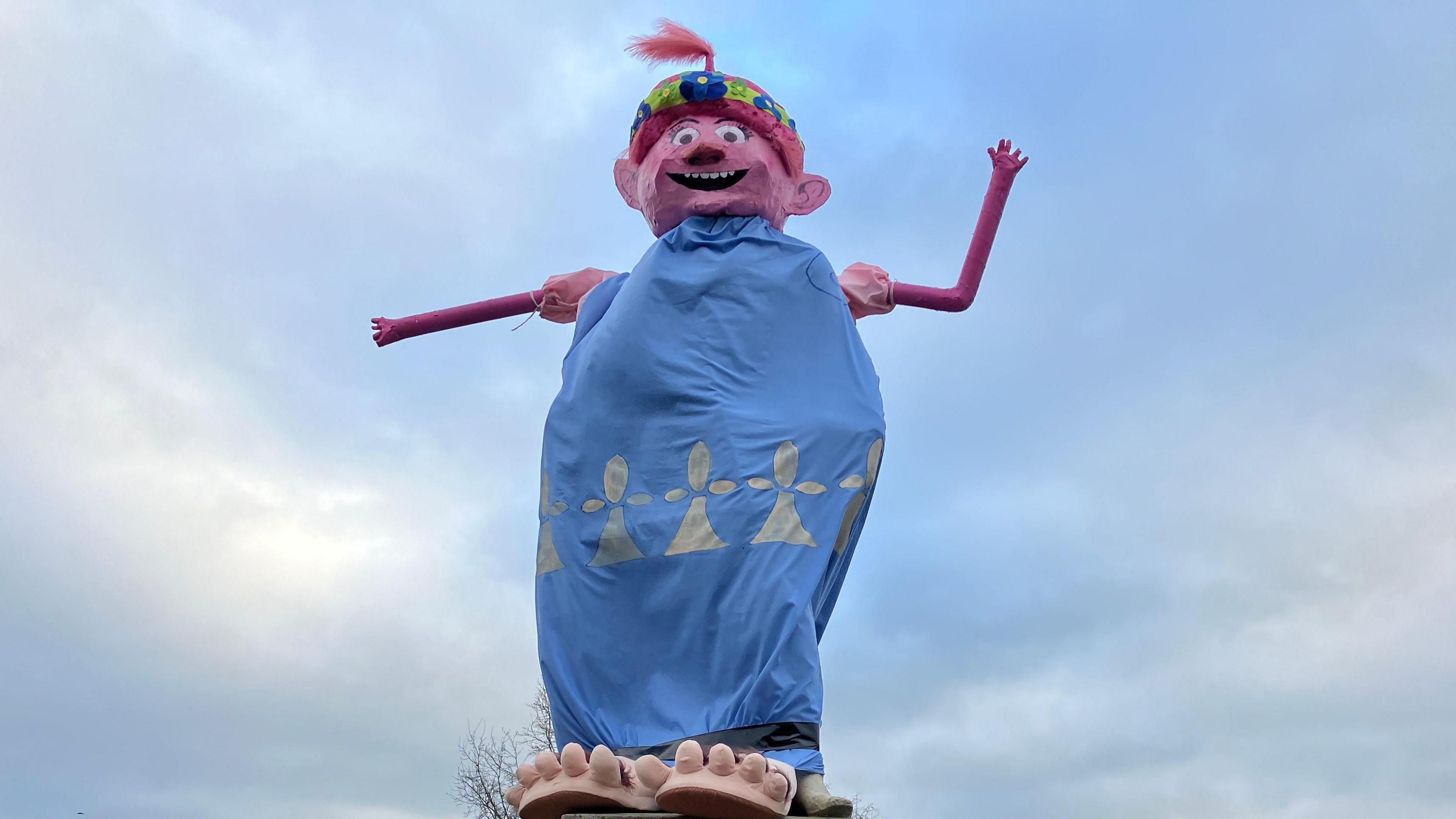 Statue decorated with a blue sheet-like costume topped with a bright pink head and colourful headband or turban.