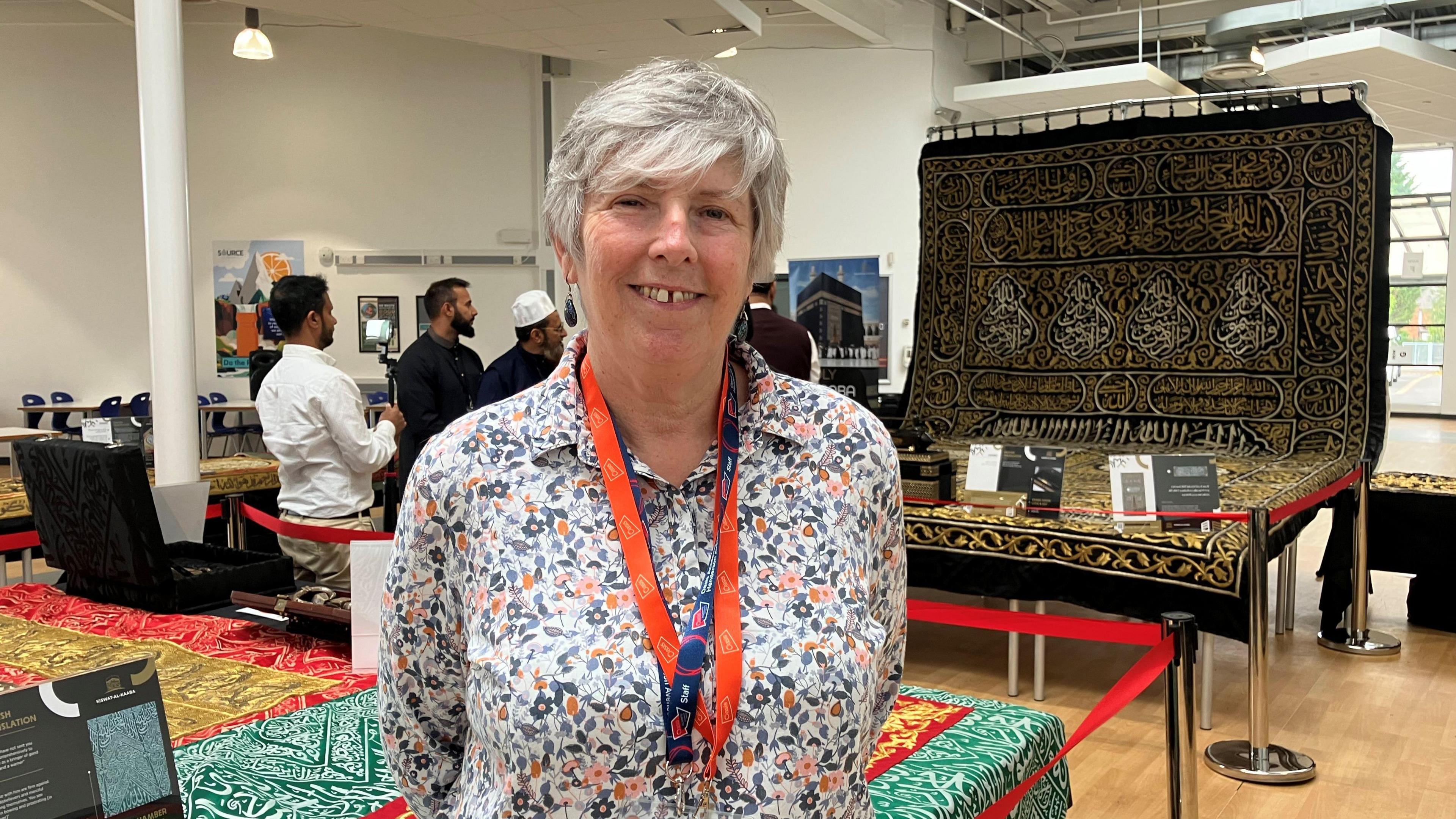 A woman with short hair, a lanyard and a patterned blouse. Behind her there are people looking at rare Islamic artefacts in a school hall.