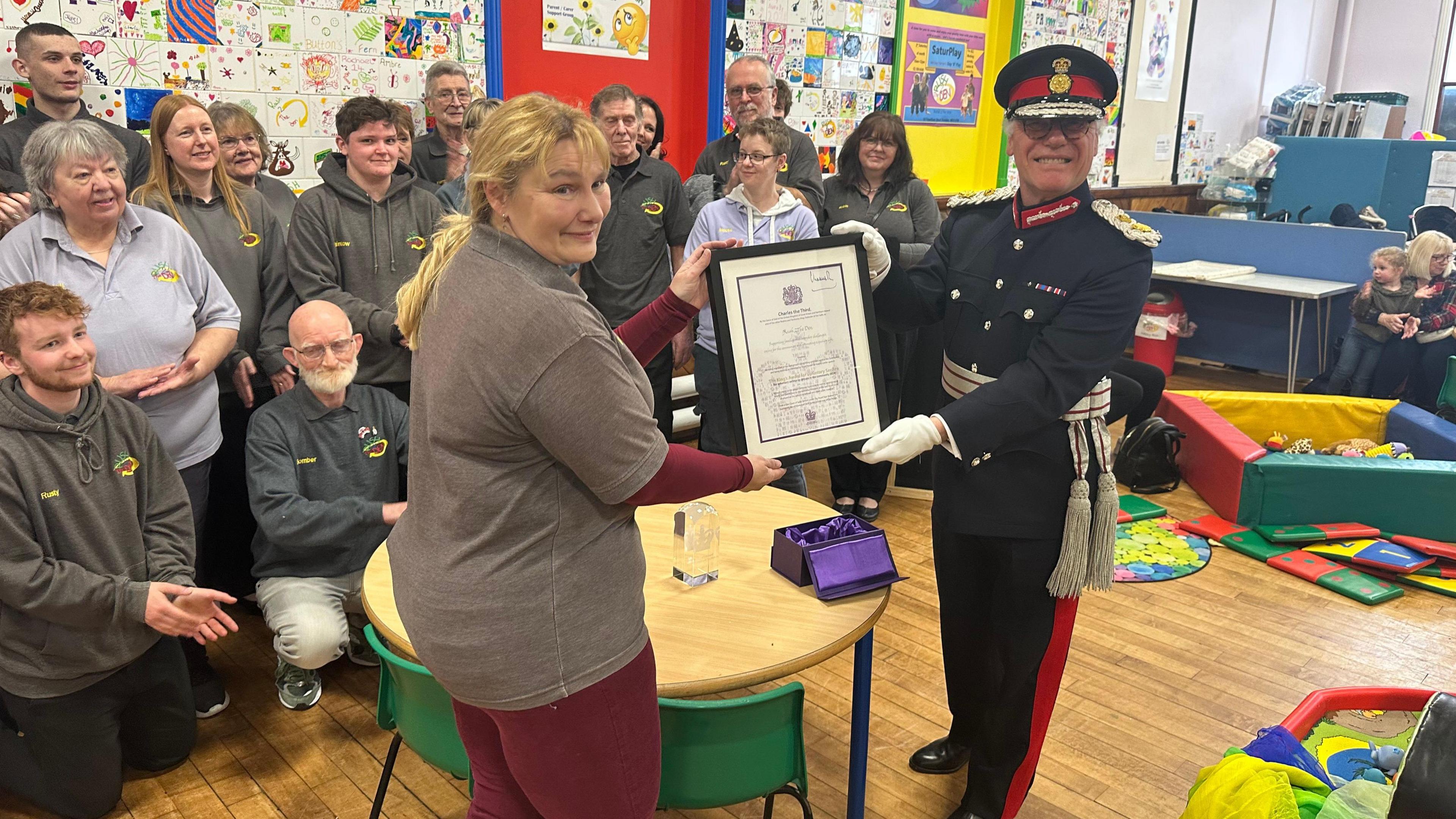 A woman in a grey sweater holds a framed award with a man in a black military uniform while a crowd of people look on.