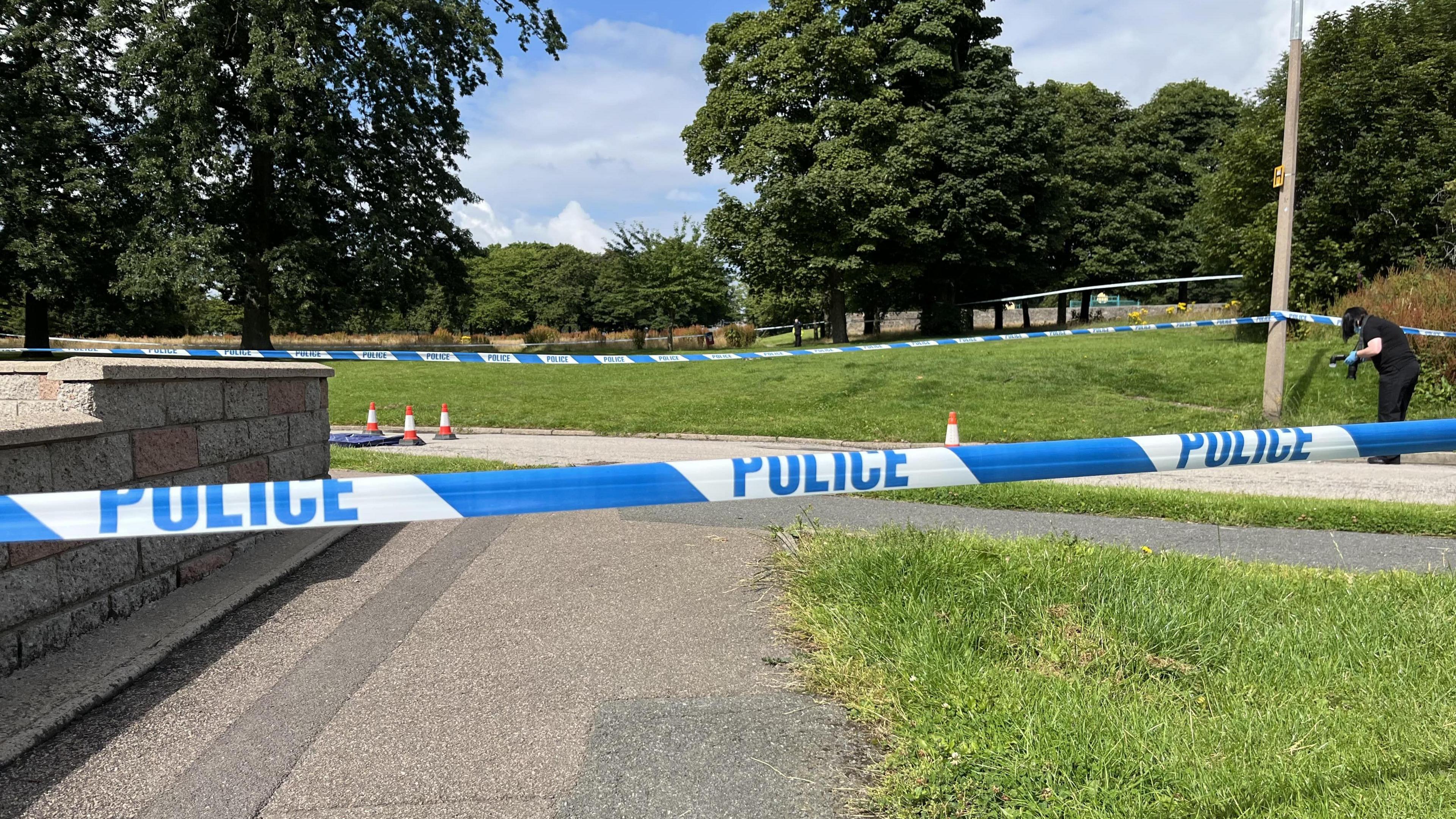 A strip of blue and white police tape across the image. There is a grey wall on the left hand side and a grey path going through the middle. A patch of green grass is on the right hand side. On the far right of the image, a police worker with long, dark hair and wearing dark clothing and blue protective gloves is partially bent over and is holding something in their hands.