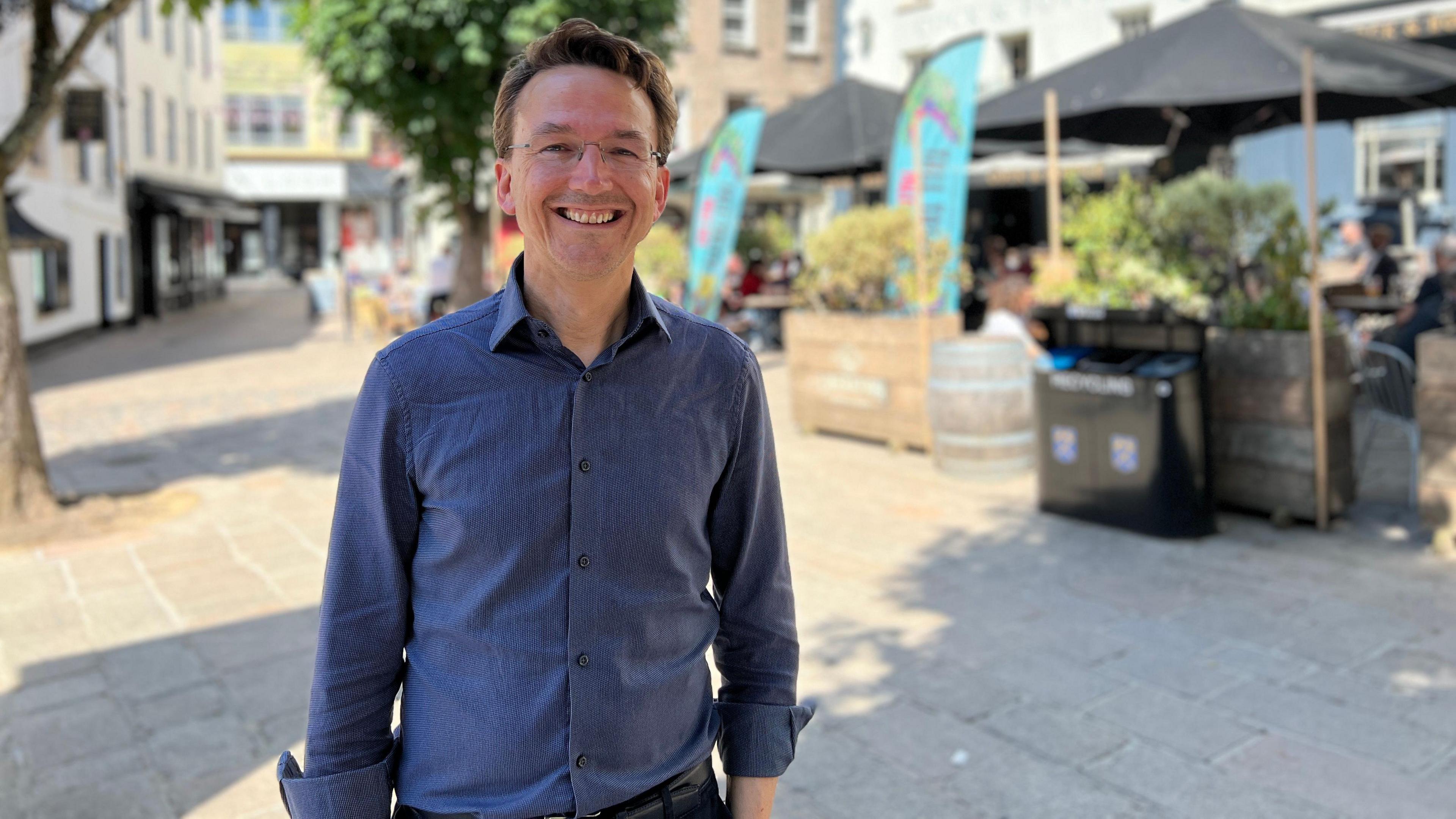 Kirsten has short brown hair and glasses and stands in an outdoor urban area on a sunny day, wearing a blue patterned shirt and dark pants. Behind them are trees, buildings, and an outdoor seating area with umbrellas and people at tables