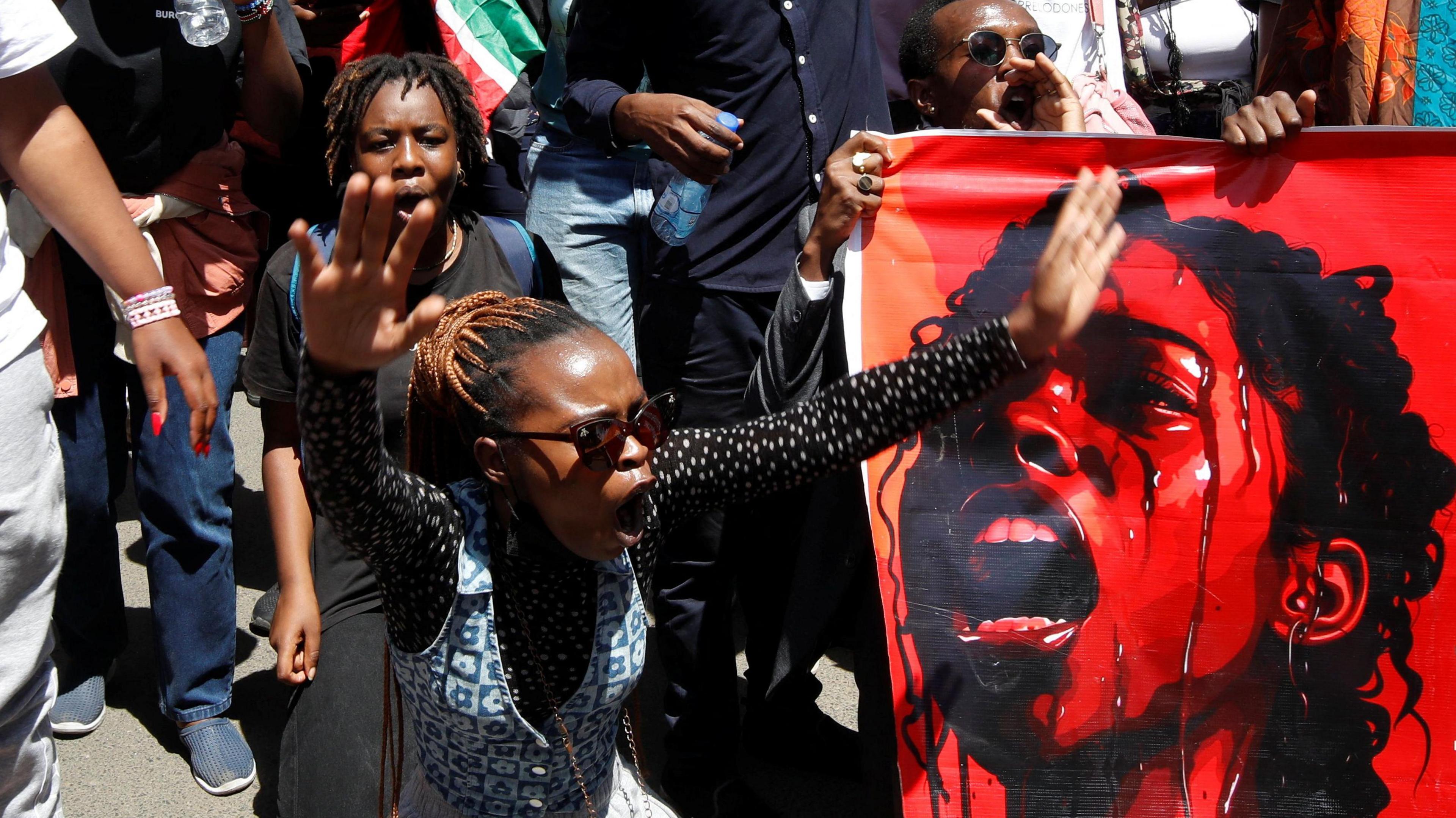 Protesters shout - one with their hands up in the air - next to a red poster showing a woman crying, Nairobi, Kenya - Tuesday 10 December 2024