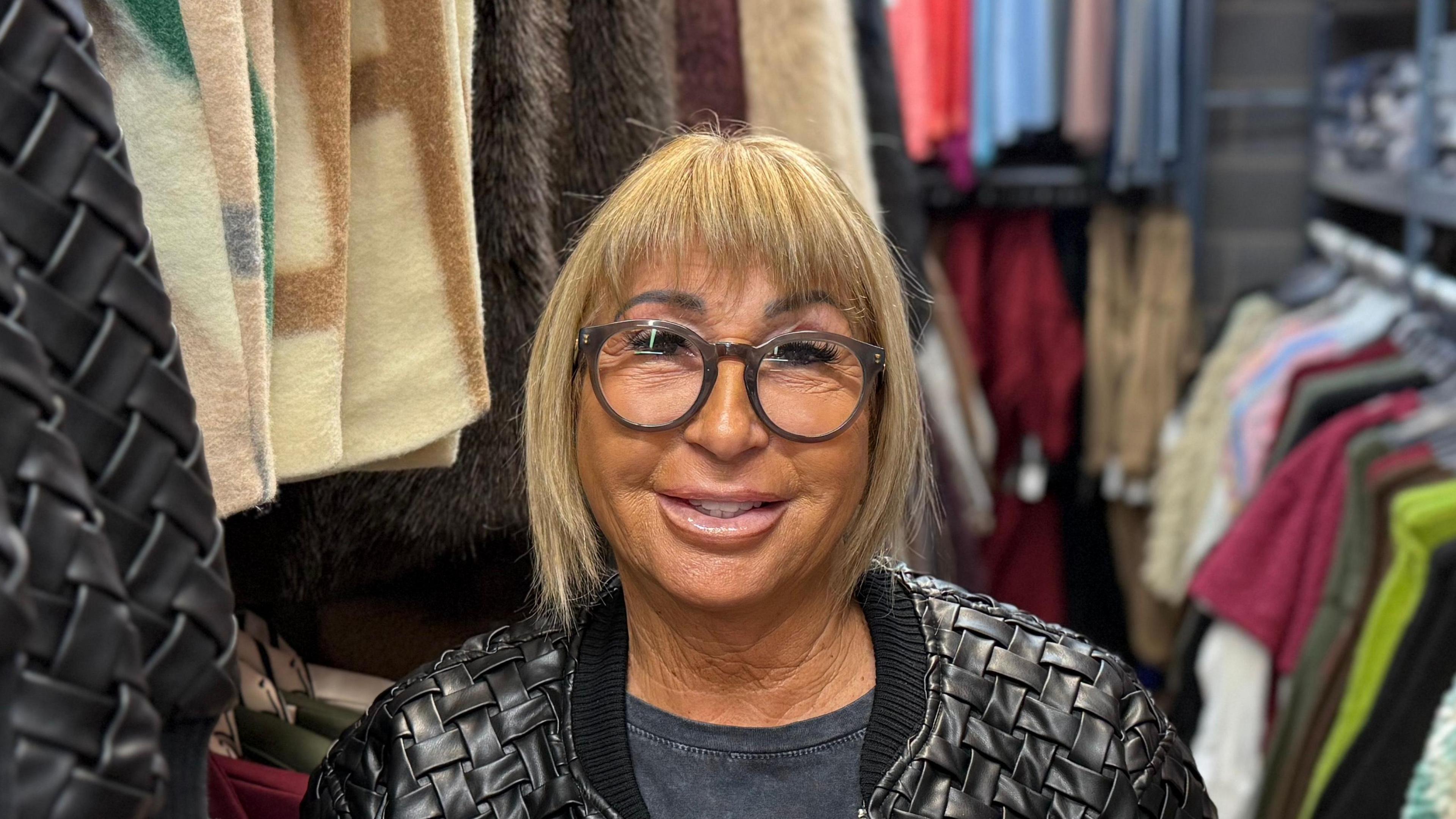 A woman - Linda Hunter - smiling straight at the camera. She is wearing a black leather jacket and dark grey T-shirt and is standing in a warehouse surrounded by clothes.