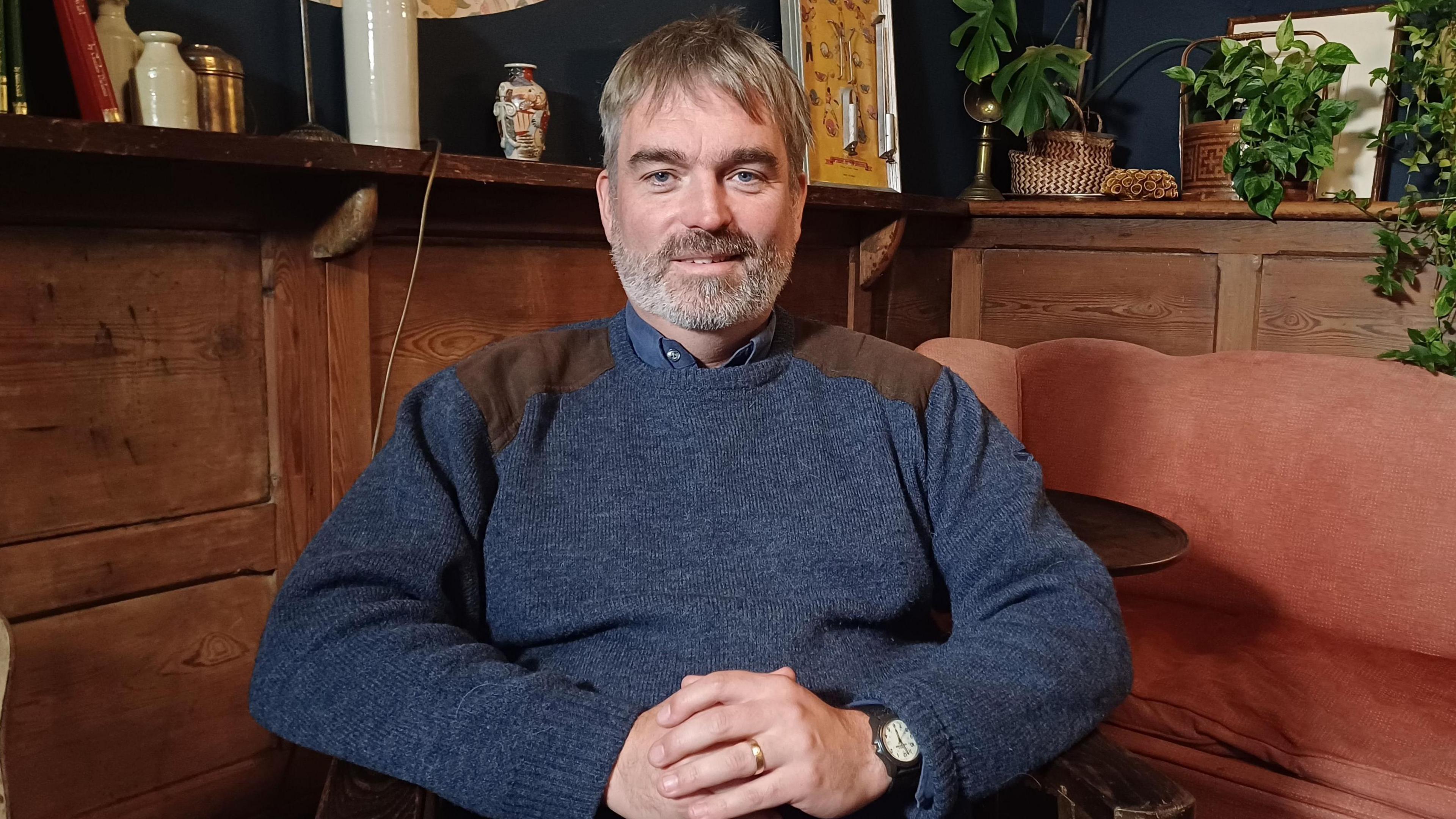 Alasdair Satchel has fair hair and a beard. He is wearing a dark blue jumper with brown patches on the shoulders. He has his hands folded in his lap as he sits in a chair in a room with wooden furnishings.