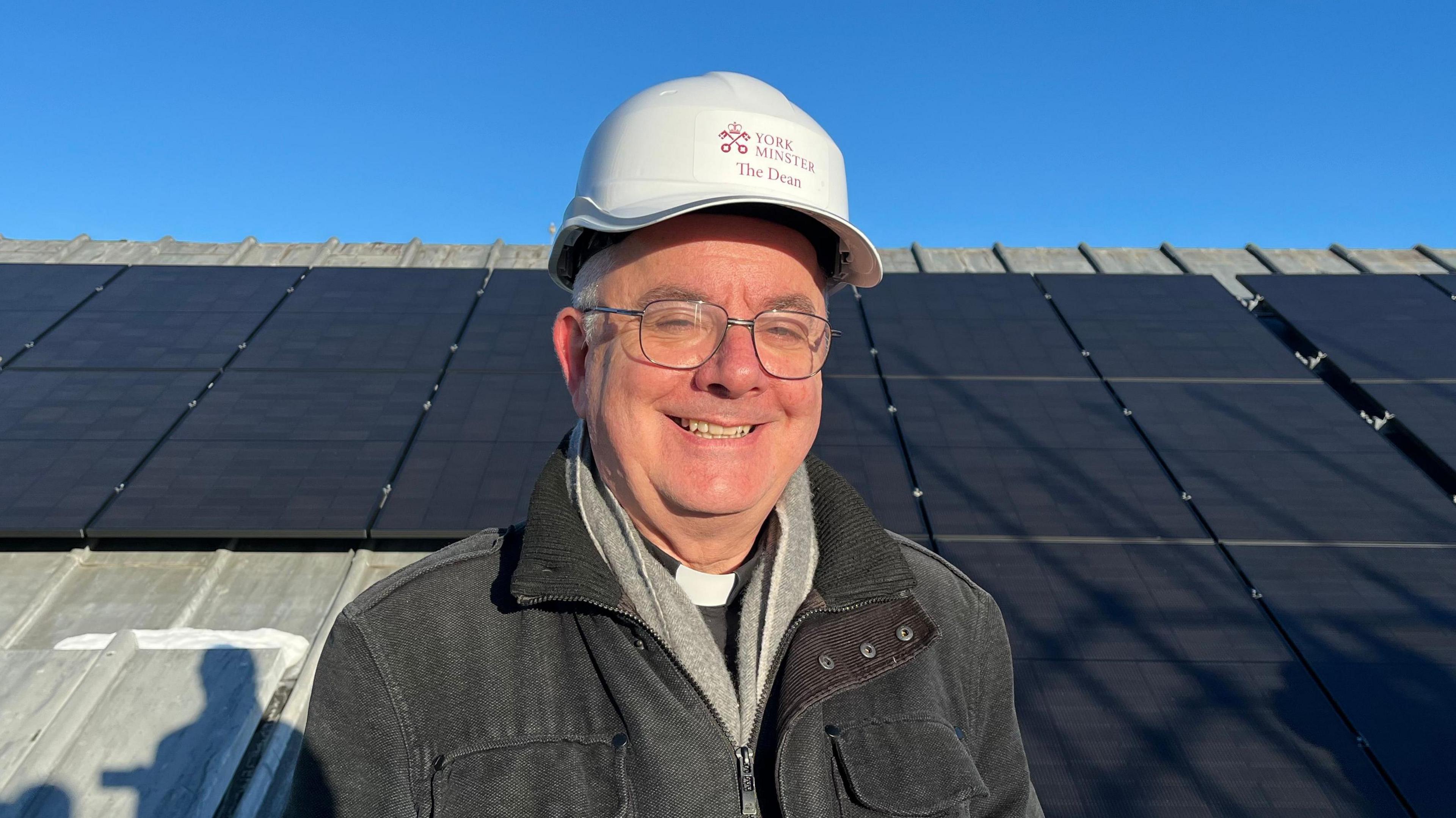 A man in a white hard hat is standing in front of several solar panels. He is wearing glasses, a black jacket, a grey scarf and a white dog collar.
