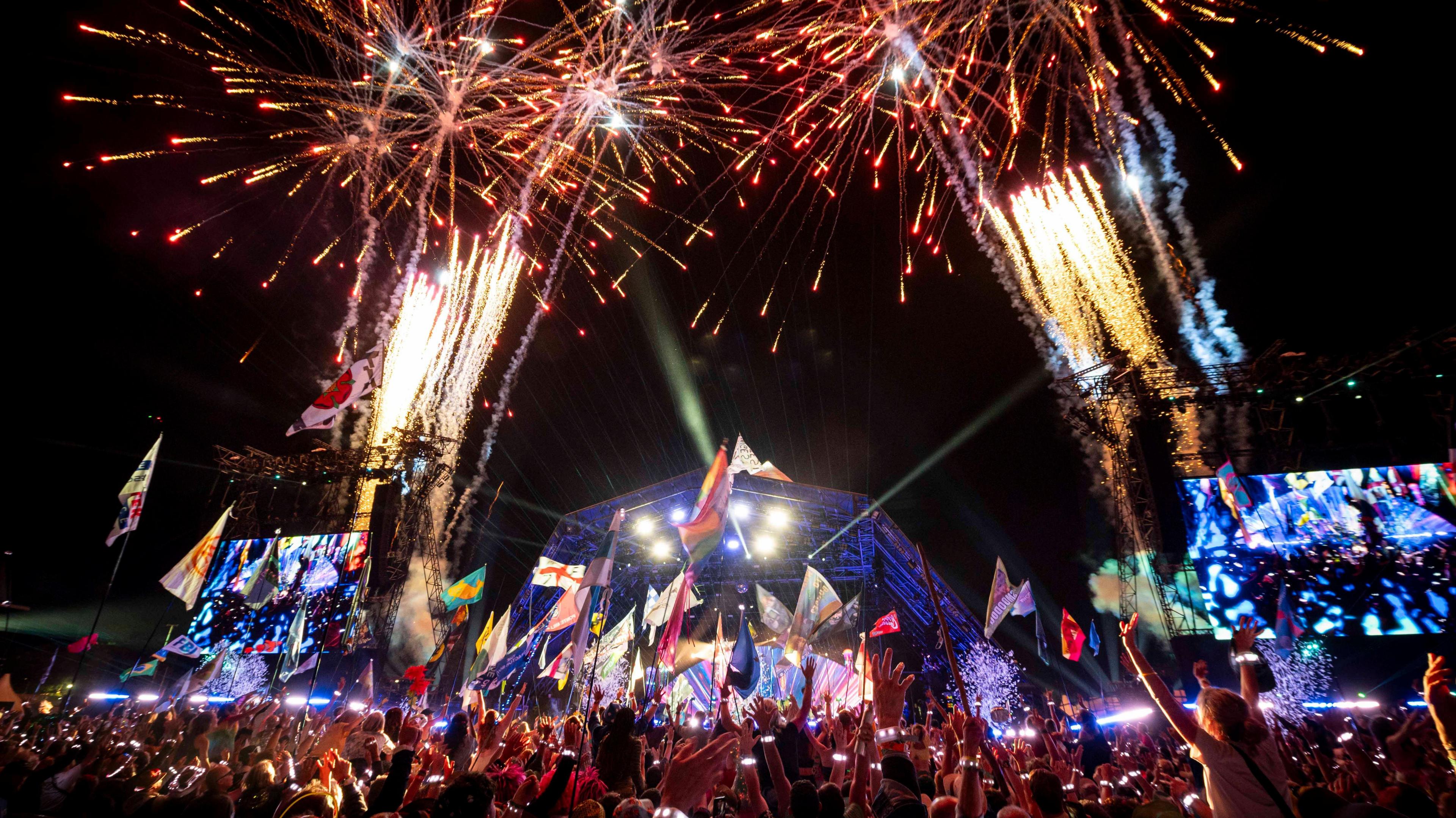 A crowd of people at Glastonbury's pyramid stage. People are holding flags and there are fireworks in the air.