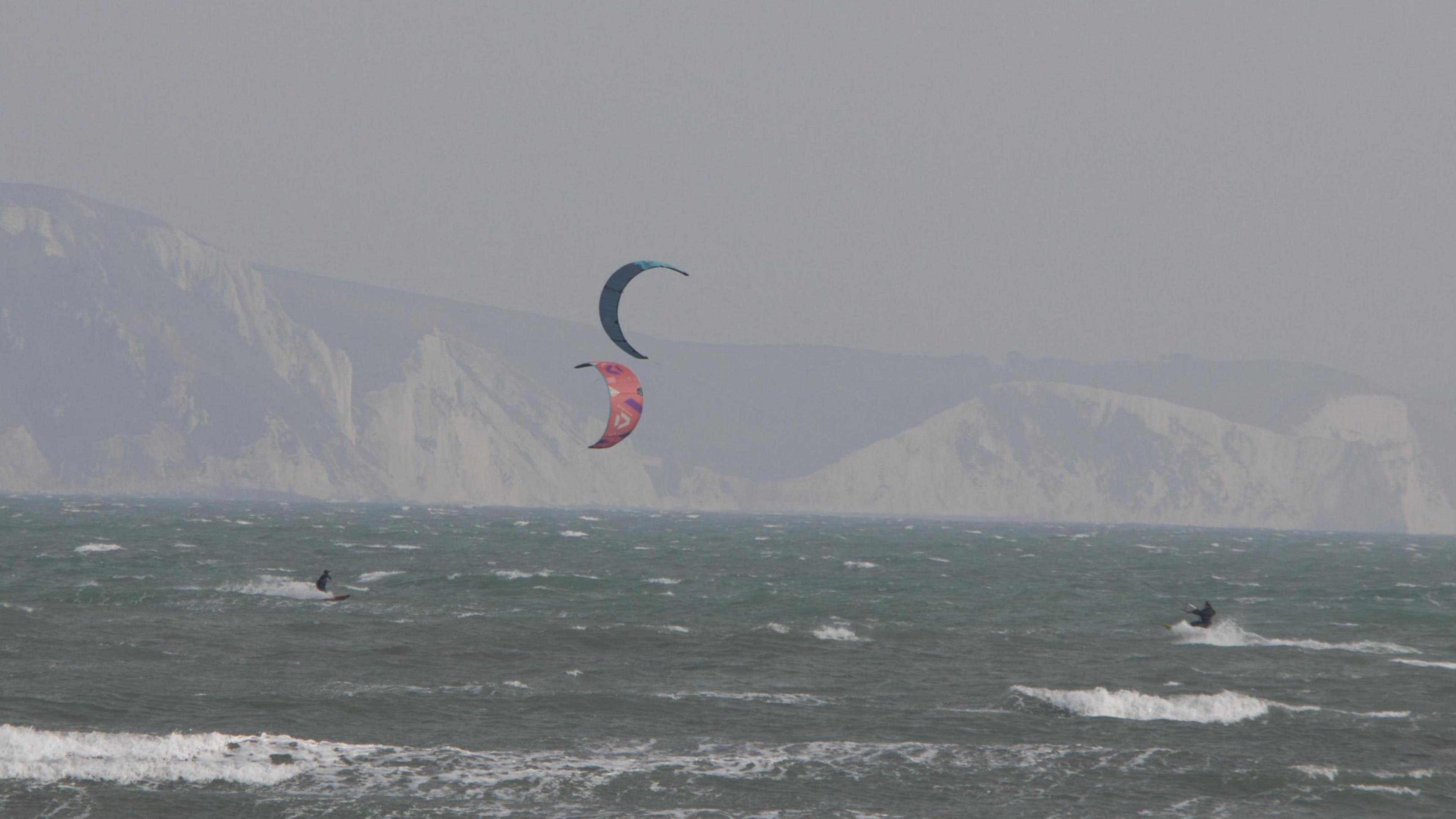 Two kitesurfers in a rough sea, with the coastal cliffs in the background. One kite is mainly red, the other blue.