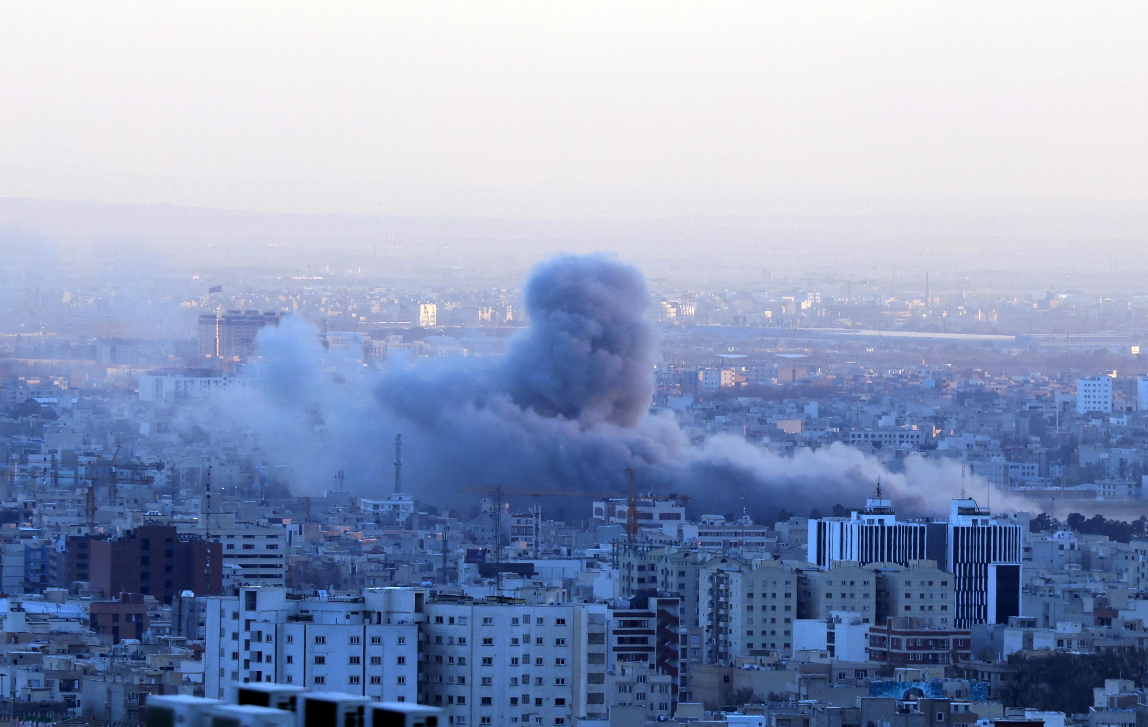 Aerial view over a cityscape. A large cloud of grey smoke rises above buildings.