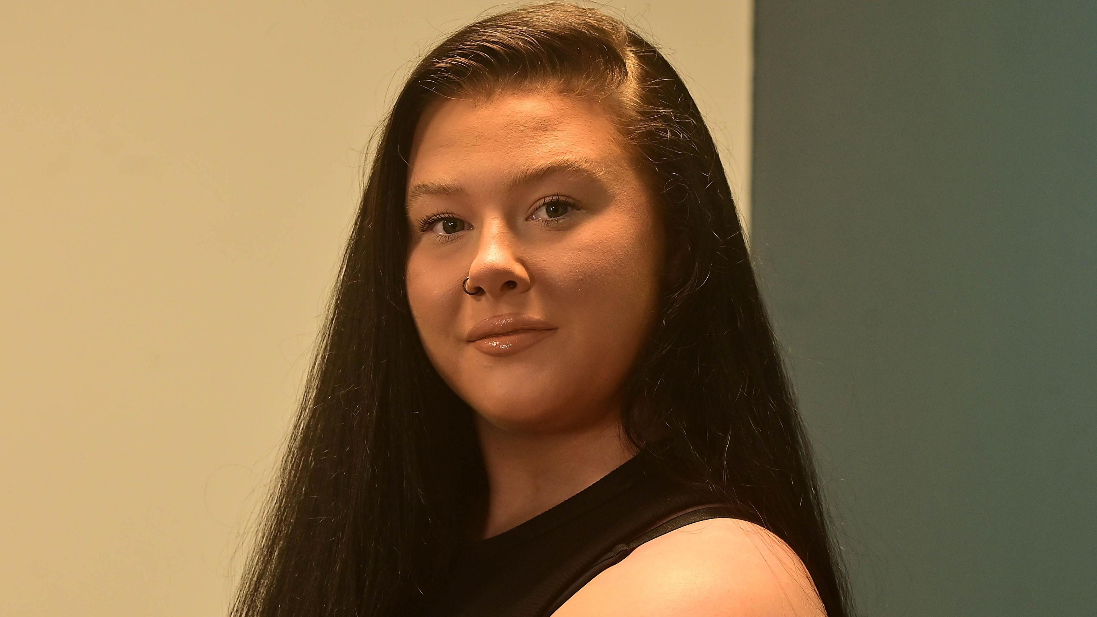 Ms Storey is wearing a black top. She's standing in front of a white and green wall. She has long brown hair.