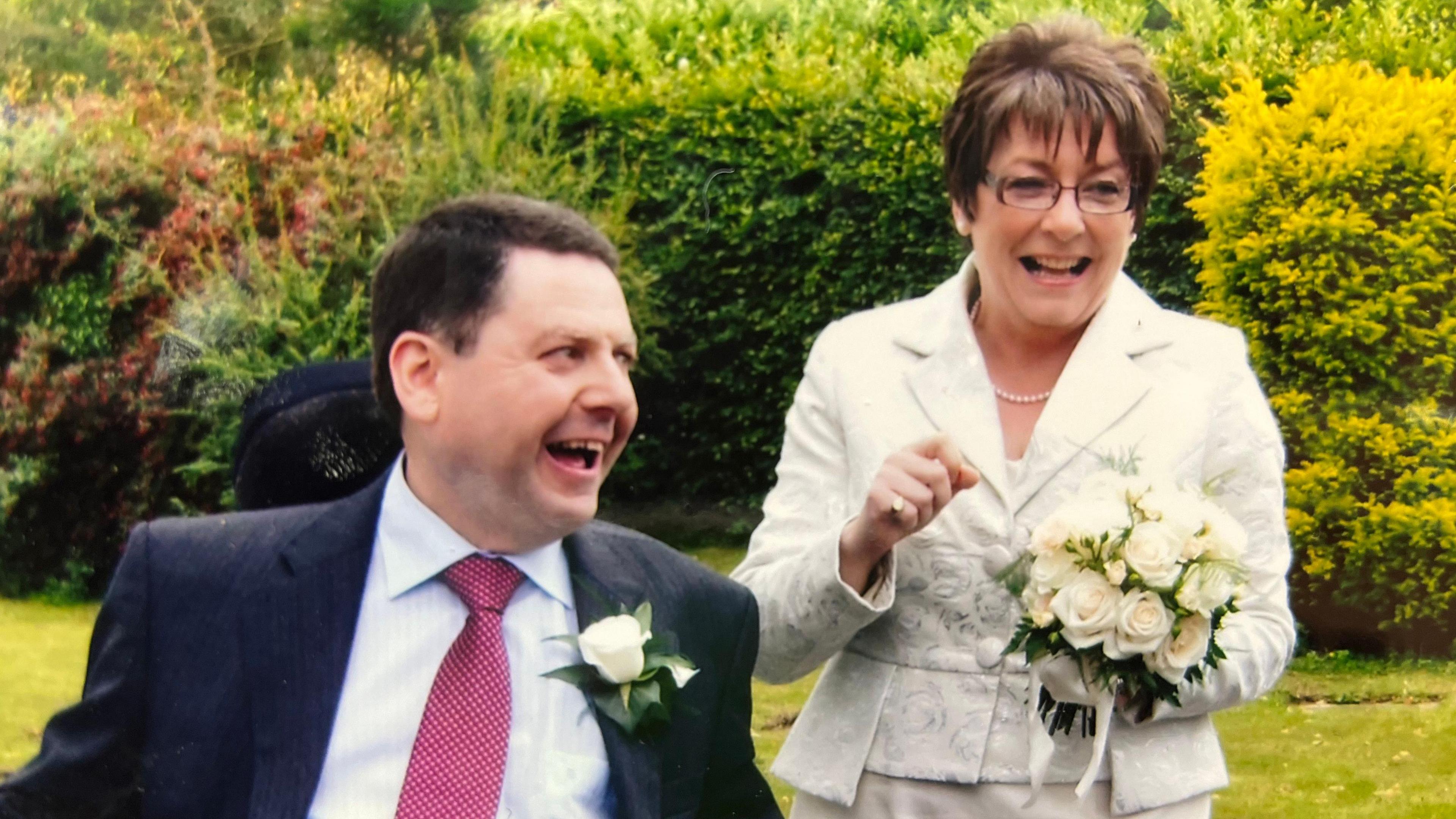A photograph of the couple on their wedding day - they're both smiling and looking off towards the right of the image. John, on the left, is wearing a dark suit with a light blue shirt and red tie, and has a white rose in his buttonhole. He's using a motorised wheelchair. Catherine, on the right, is wearing a white floral suit and is holding a bouquet of white roses. They're stood outside, with grass and a hedge behind them.