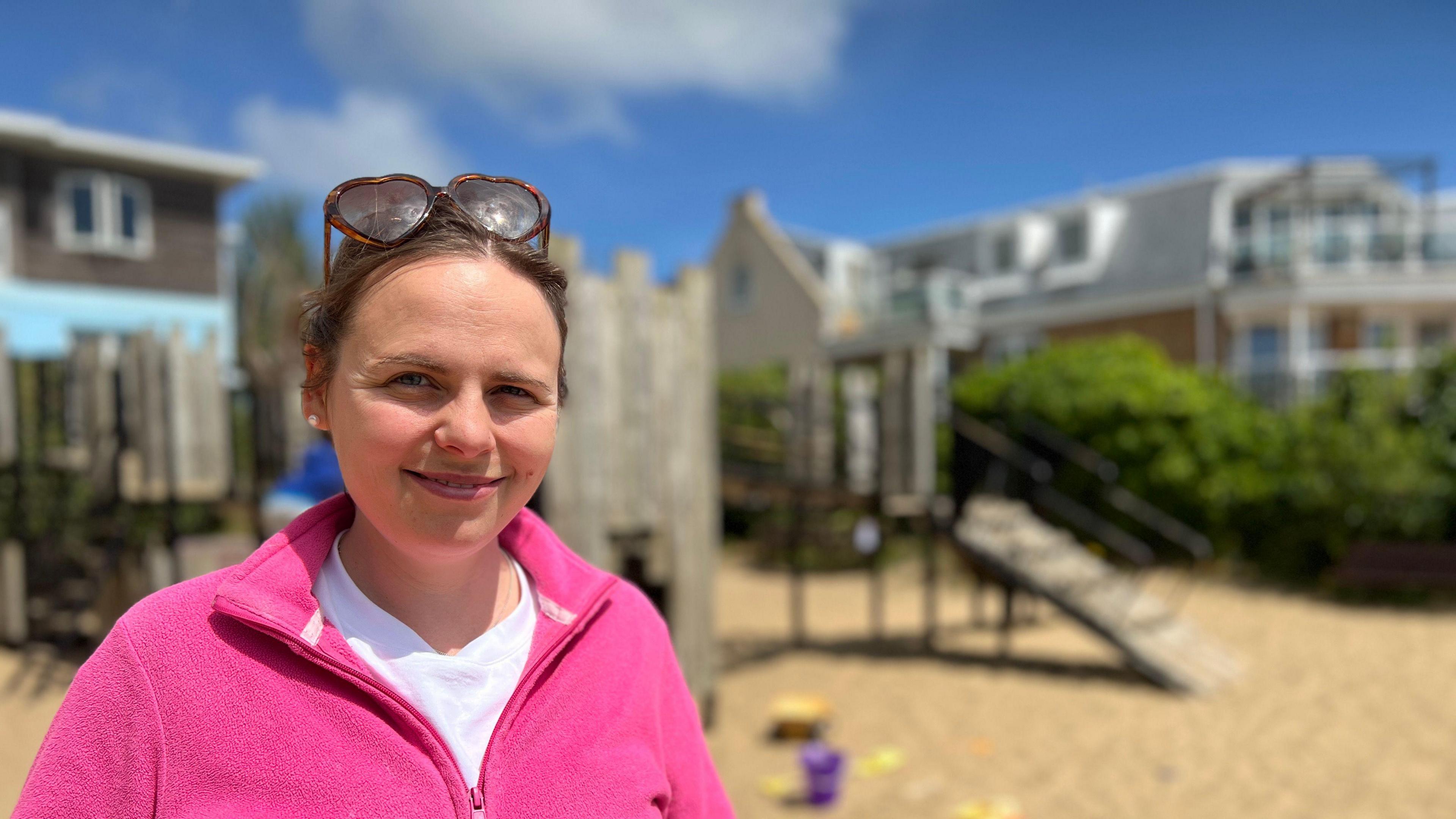 Charlotte stands smiling at the camera. The play park is visible in the background. She is wearing a pink fleece, has brown hair and has a pair of heart-shaped sunglasses on her head.