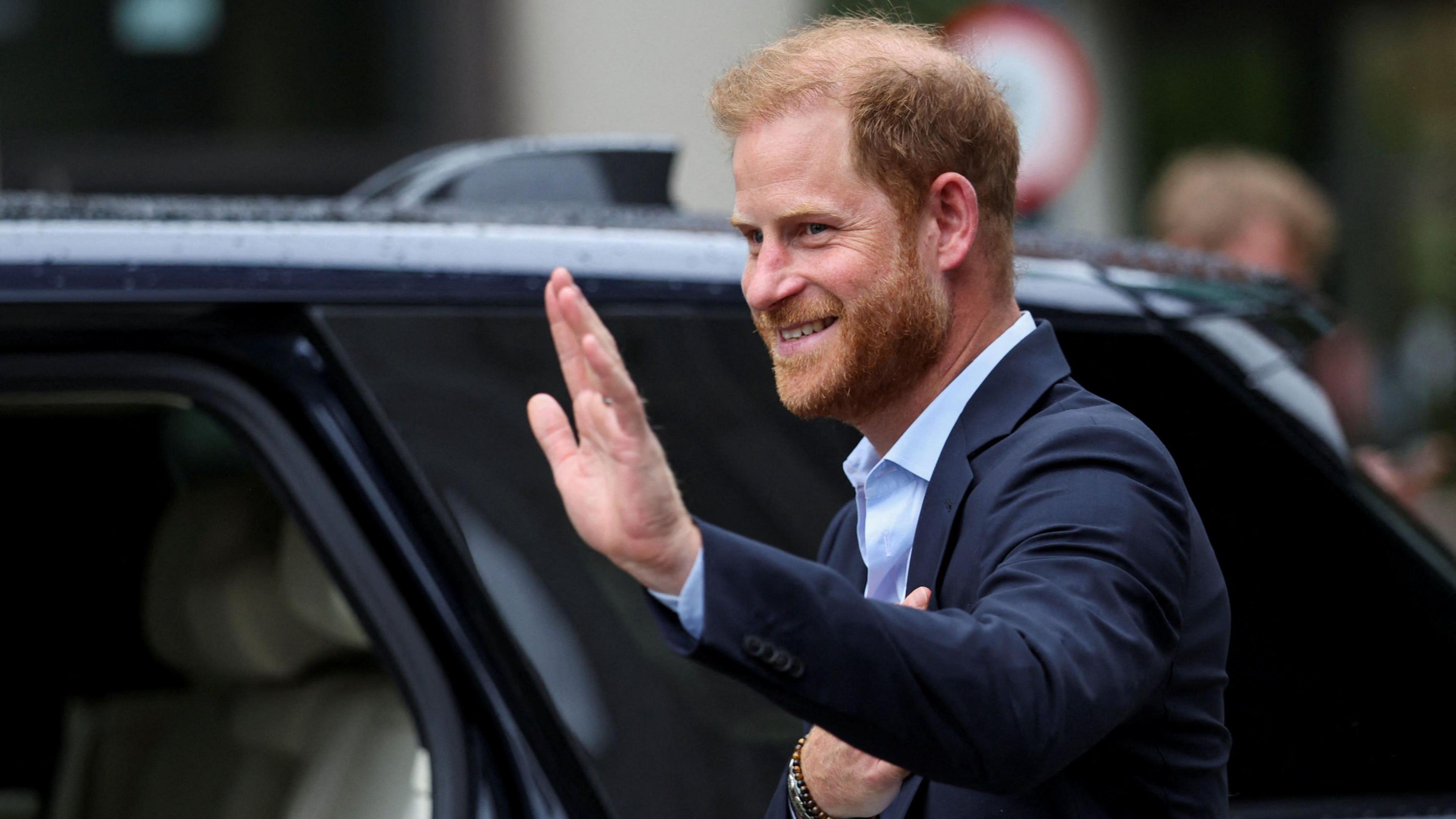 Prince Harry wearing a blue suit and blue shirt, waving as he steps into a black car.