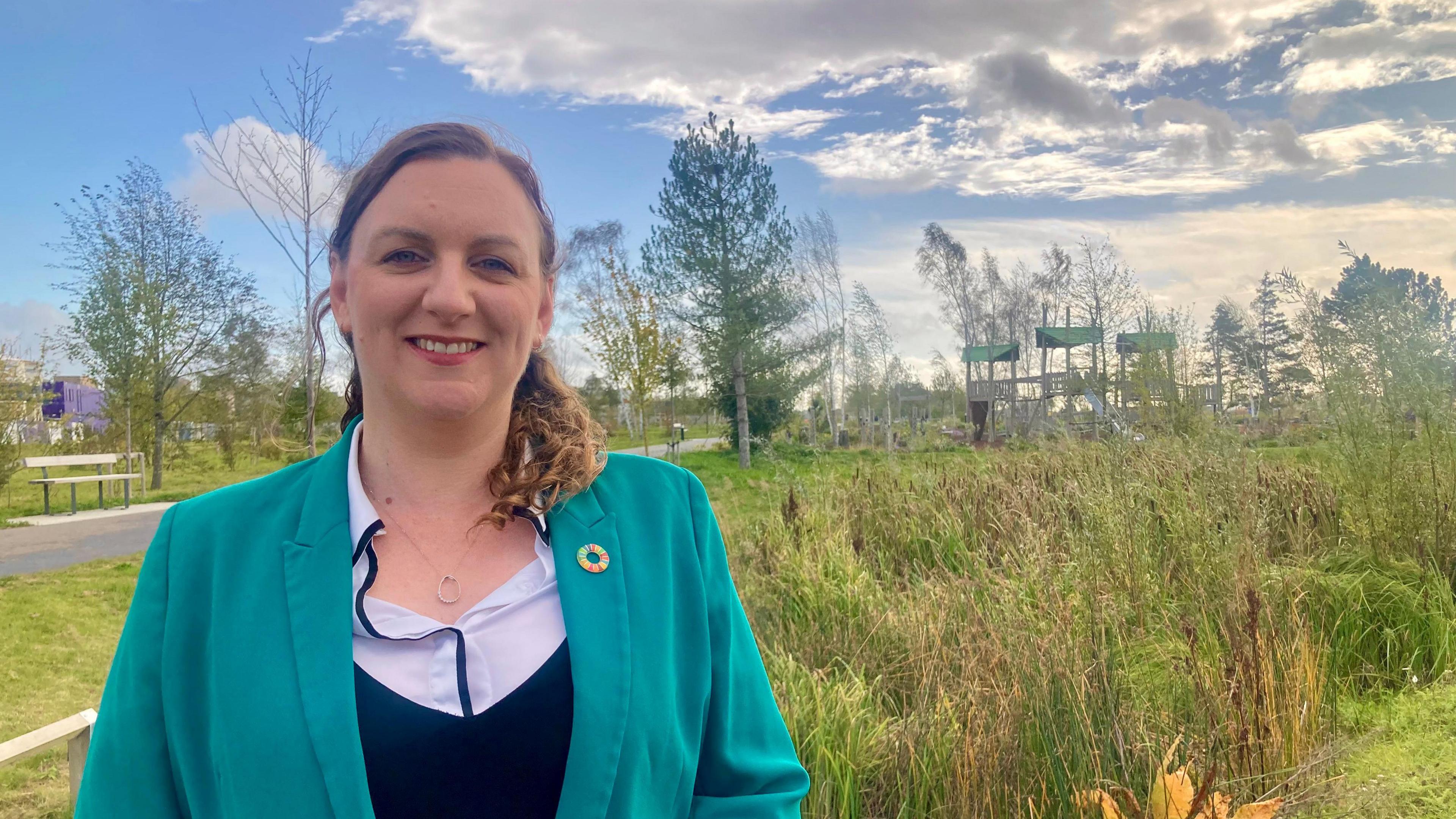 Elizabeth Orchard is smiling at the camera, set to the left of the photo, and is wearing a green jacket with a black and white jumper underneath. She has brown shoulder-length hair tied back in a ponytail. In the background is a green space with a children's play area. It is a sunny day with some clouds in the sky. 