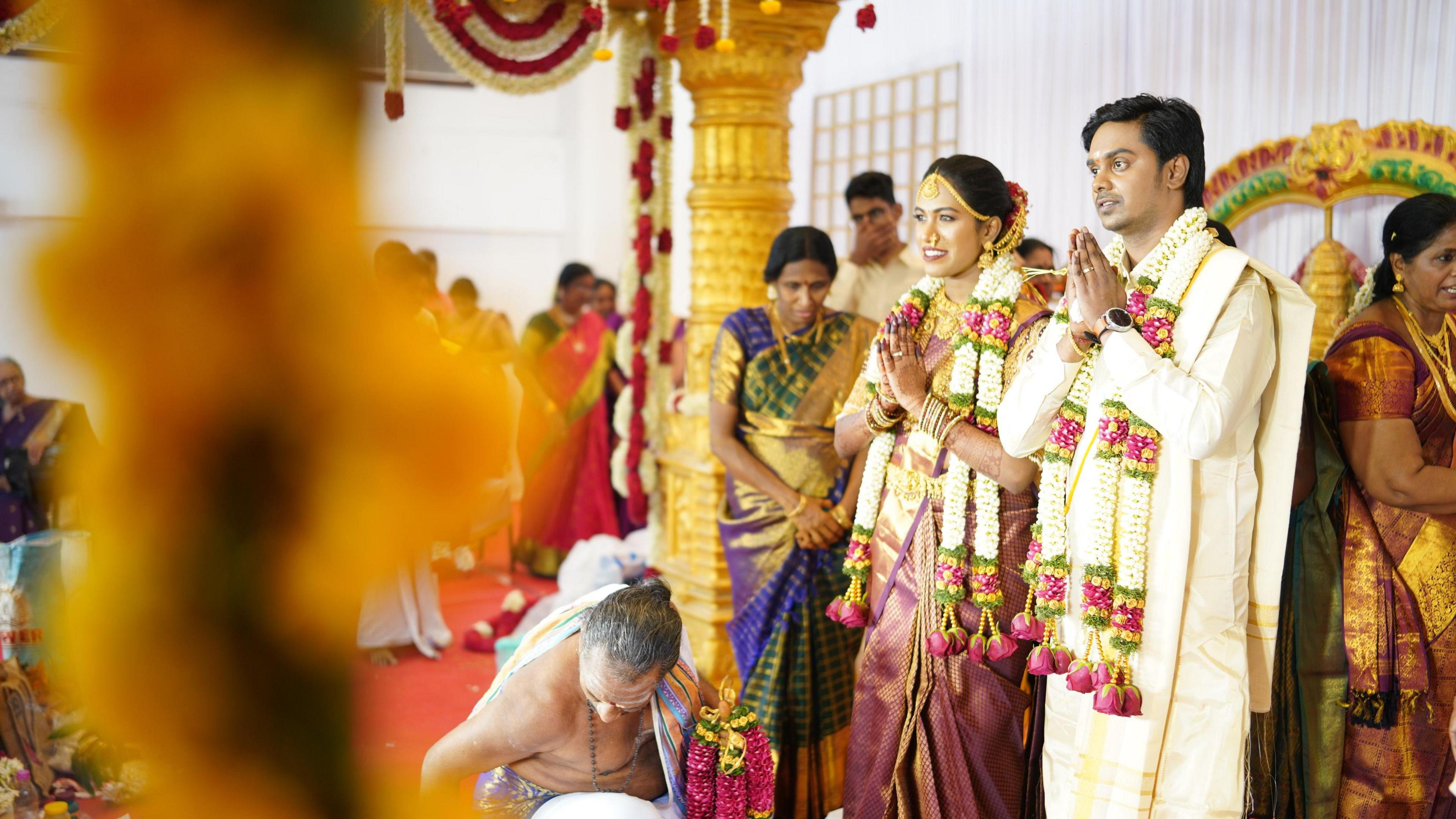 Janani Mohan and her husband in traditional Indian wedding clothing with garlands round their necks, stand with their hands in a prayer position in front of them, surrounded by guests celebrating their wedding