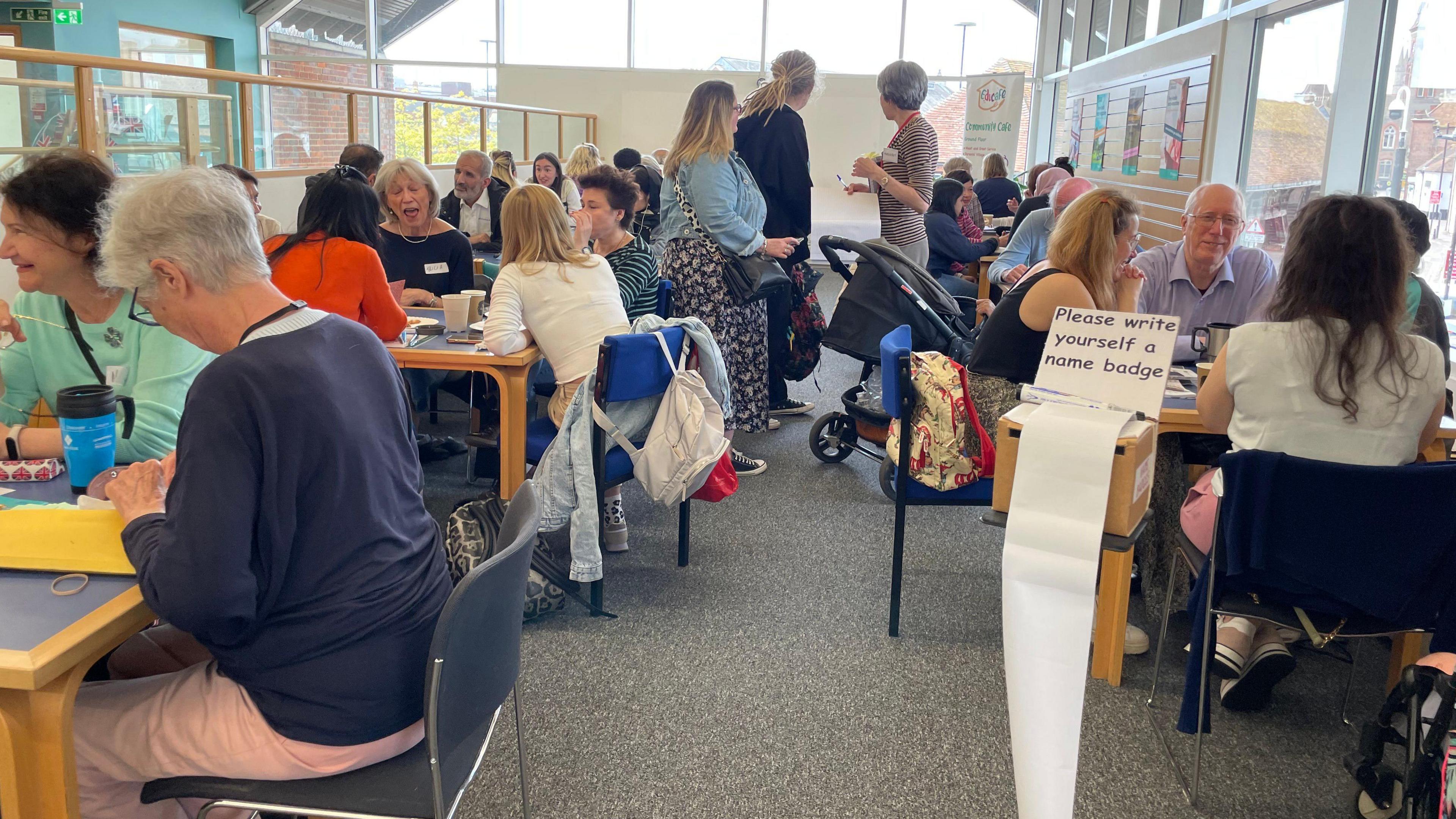 A room in a library with lots of people sitting round small tables chatting