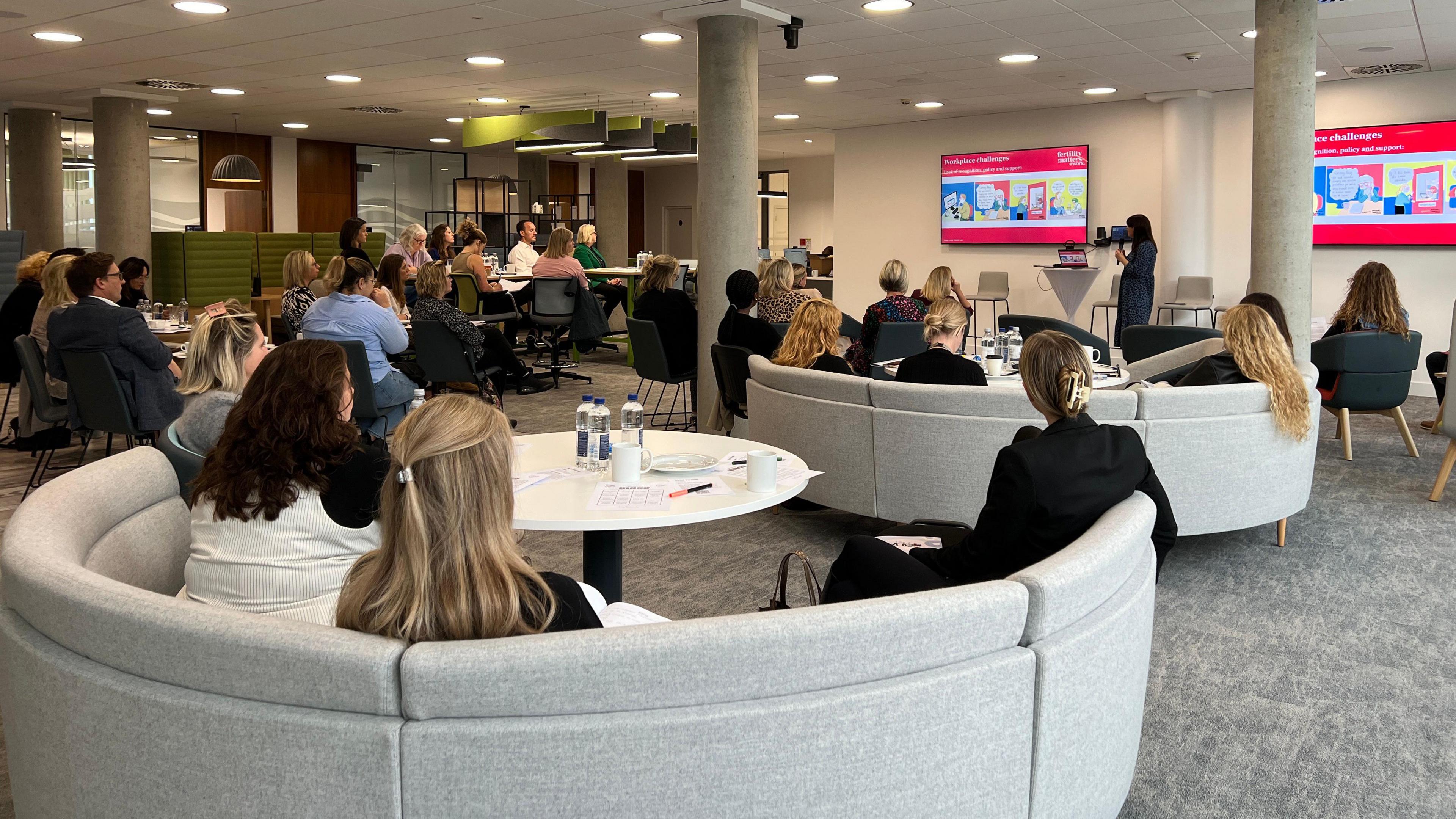 A group of people are sitting on either circular, grey sofas or black chairs and they're facing a woman giving a speech. She is looking at a TV screen with animated drawings on it. There are between twenty to thirty people in the room. There are mugs of coffee and tea on some tables along with bottles of water.