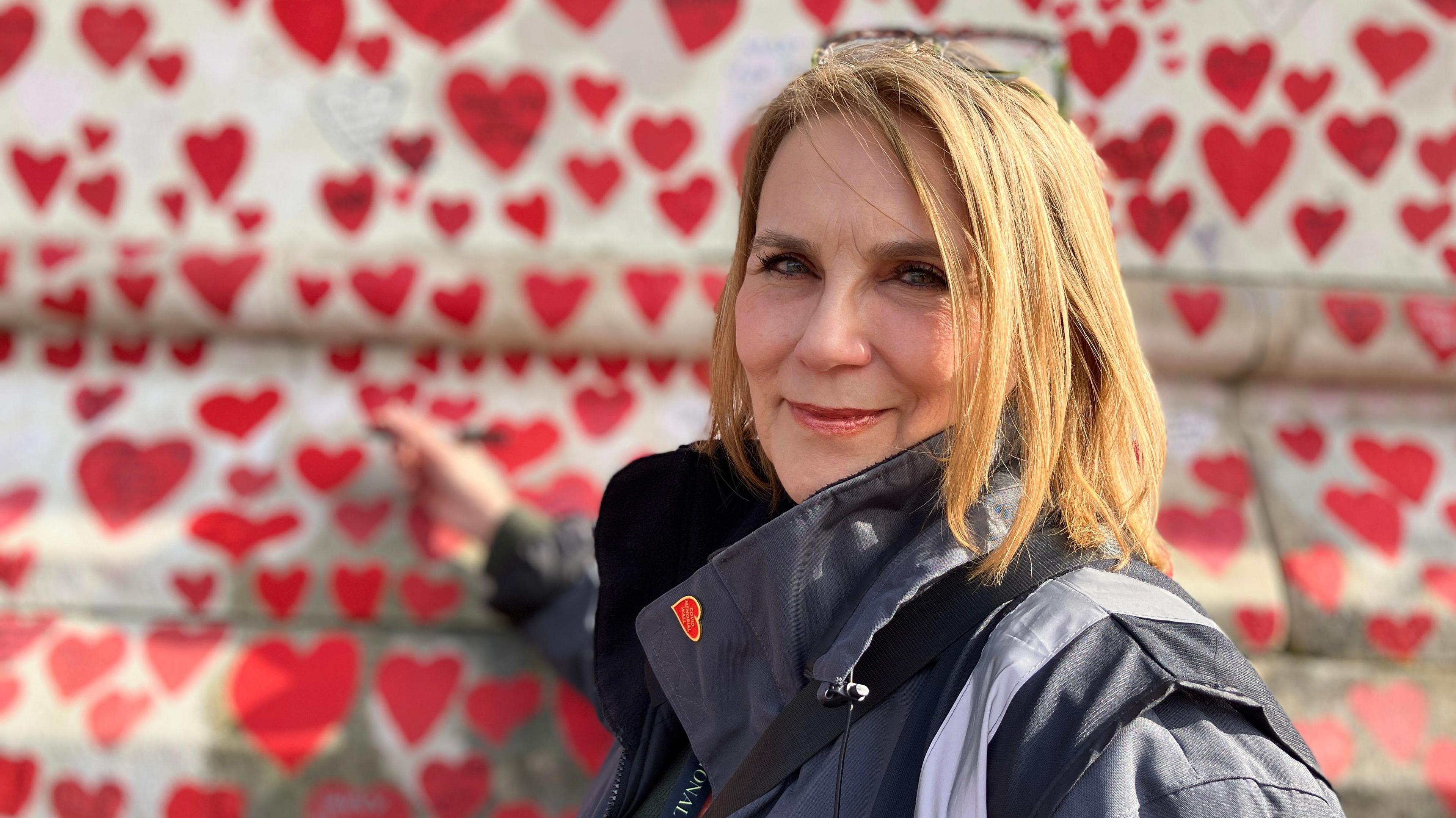 A woman with blond hair holds a pen to a wall covered in red hearts. She has on a jacket and red heart pin badge on the collar