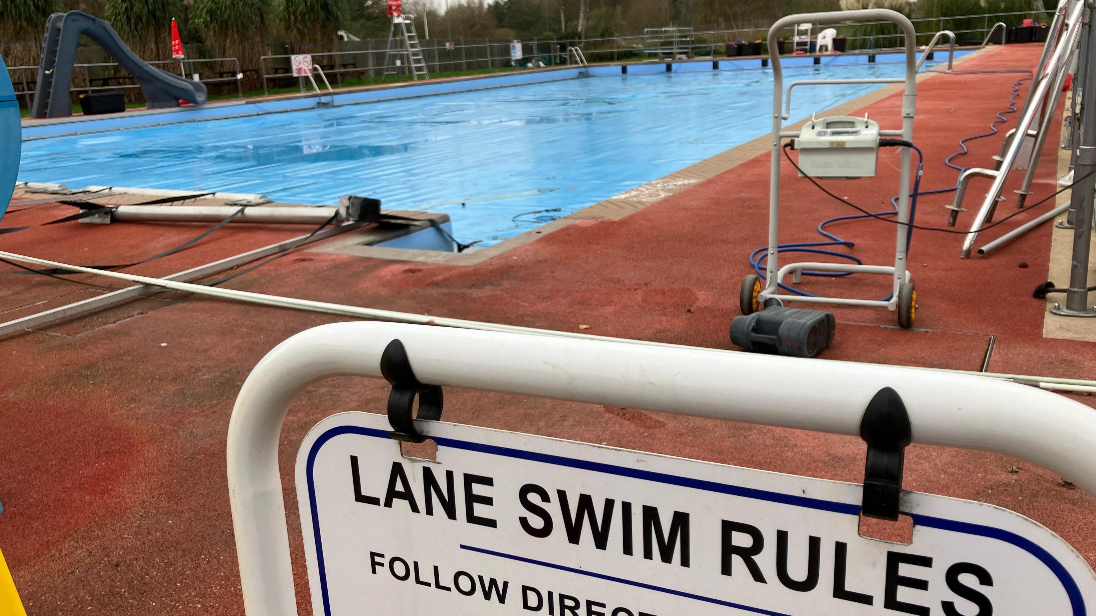 An outdoor swimming pool with a blue cover surrounded by red tarmac with a white sign in the foreground reading " Lane Swim Rules"   