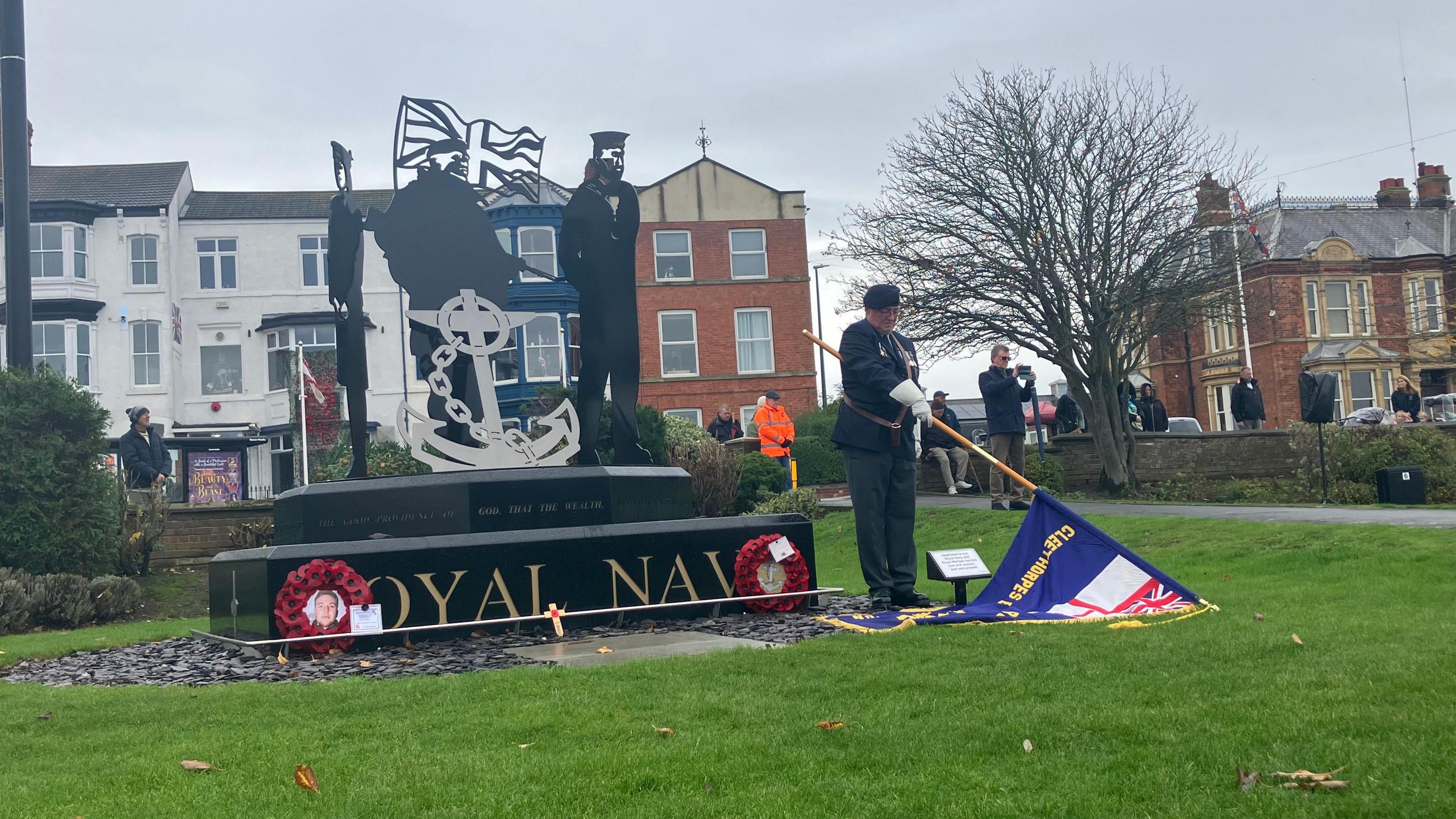 A man dressed in military uniform holding a blue flag pointed towards the floor. He is standing in front of a Royal Navy memorial, with wreaths at either side. The memorial displays crew members and an anchor.