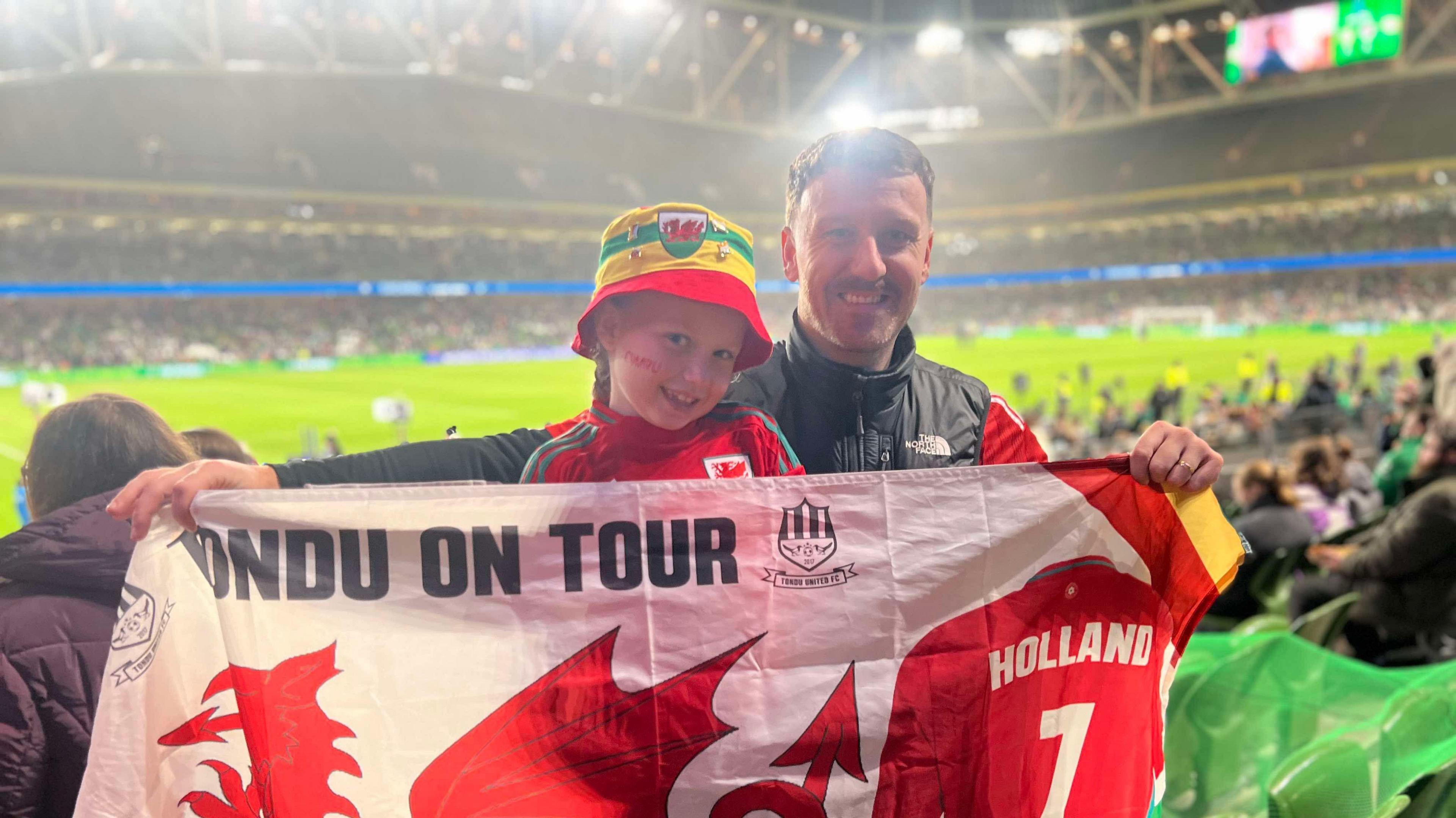 Joey Healey and Harlow supporting Wales in Dublin. They are holding a Wales flag, with the stadium behind them. Harlow has a Wales bucket hat on and a Wales football top on.