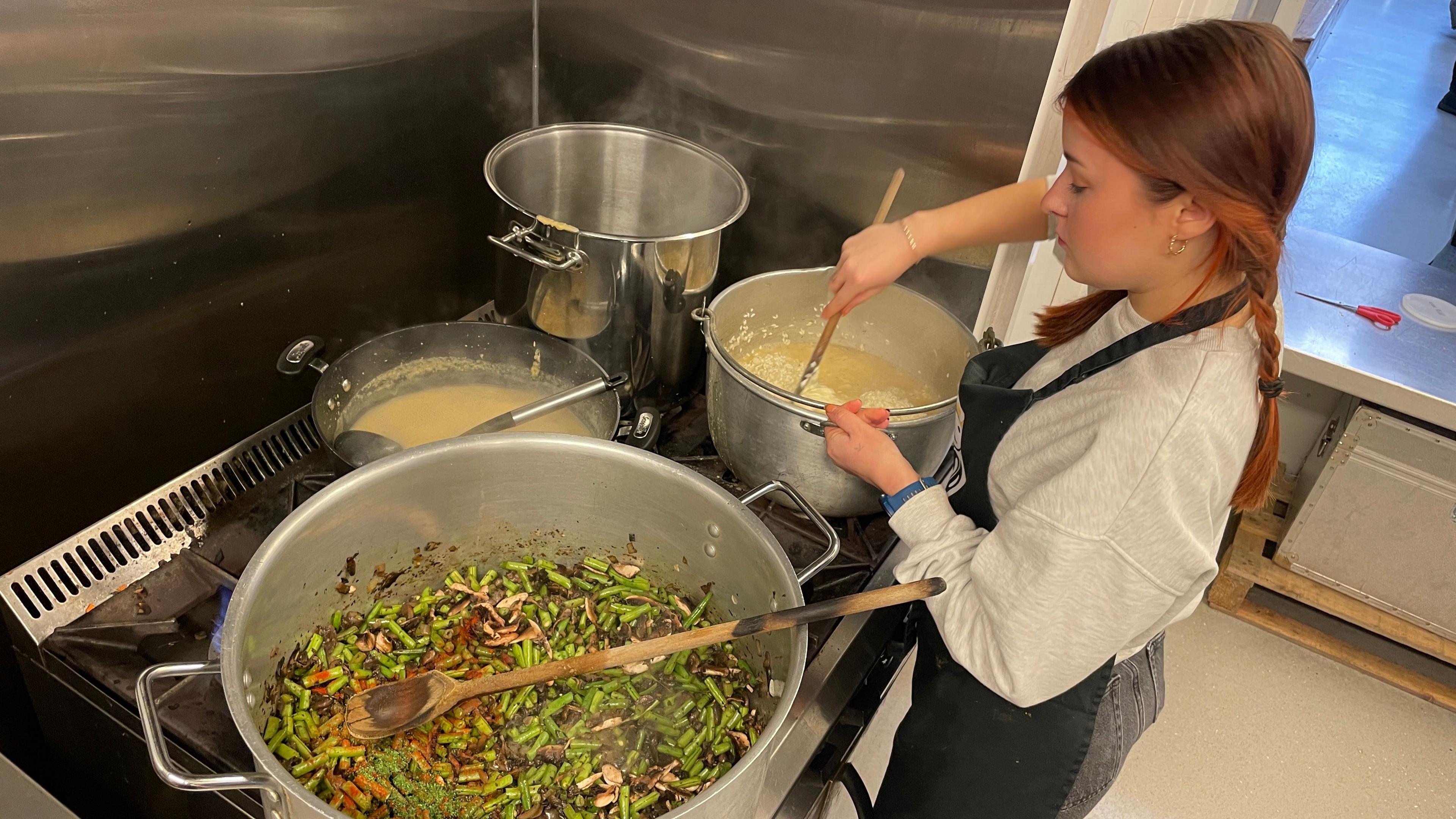 A young woman stirs some food in a metal pot on a stove. A large metal pot sits alongside the other and contains lots of green beans.