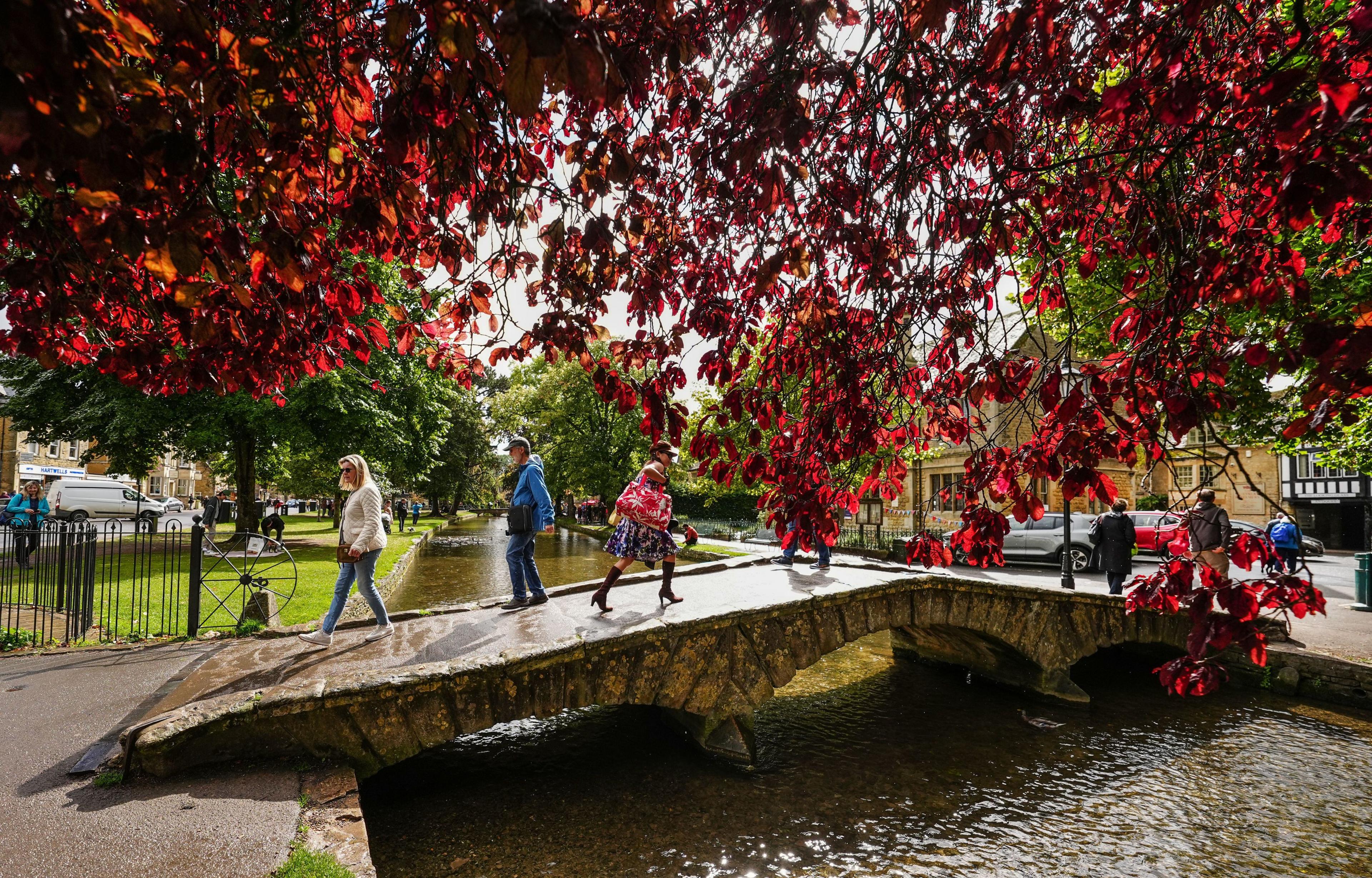 Bridge over the river Windrush in Bourton on the Water. Three people are crossing the bridge, with some red autumn leaves partly blocking the view of the river in the upper foreground. 