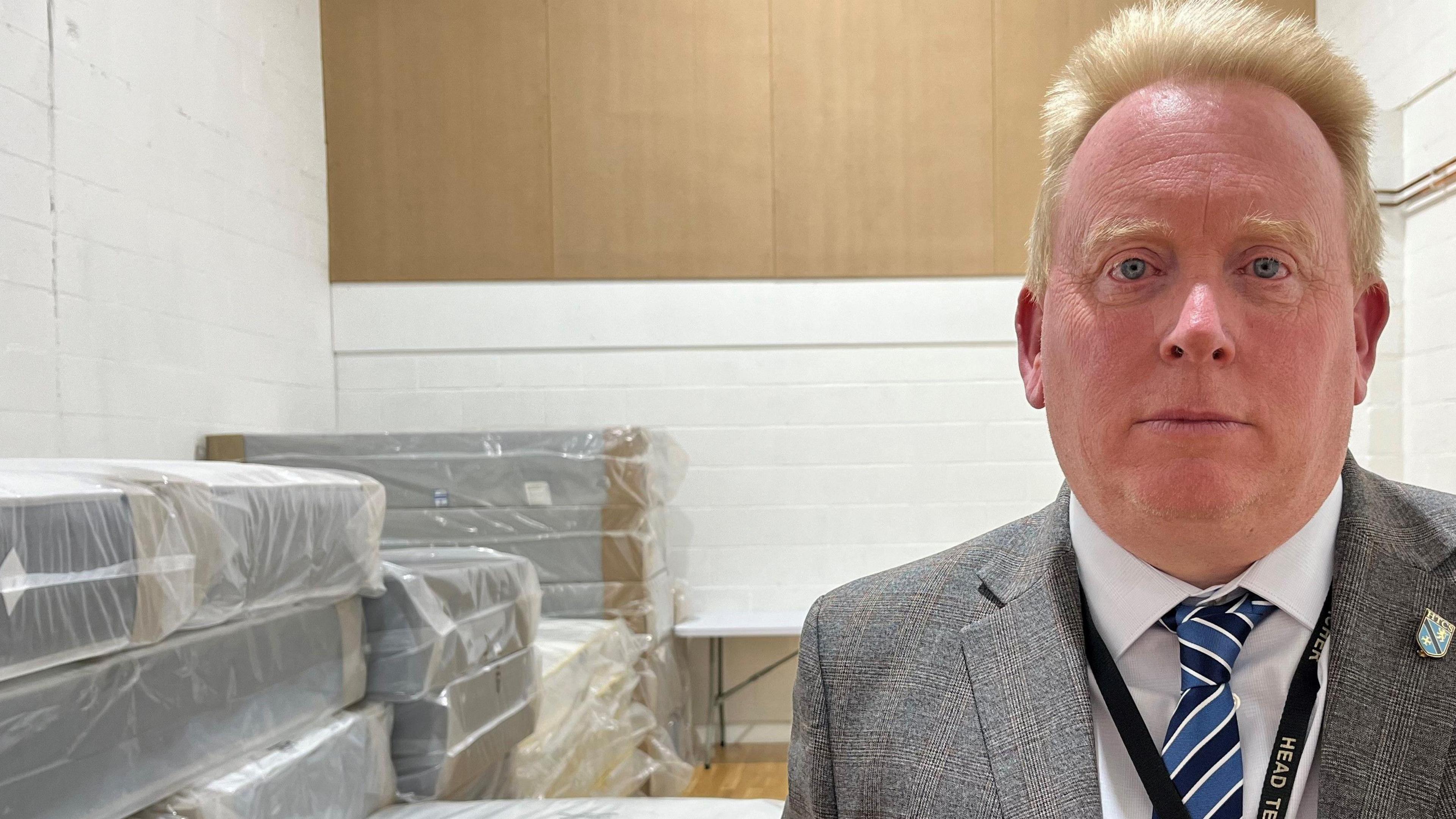 Mark Tilling, a white man with spiked blonde hair wearing a suit and tie, in front of piles of beds