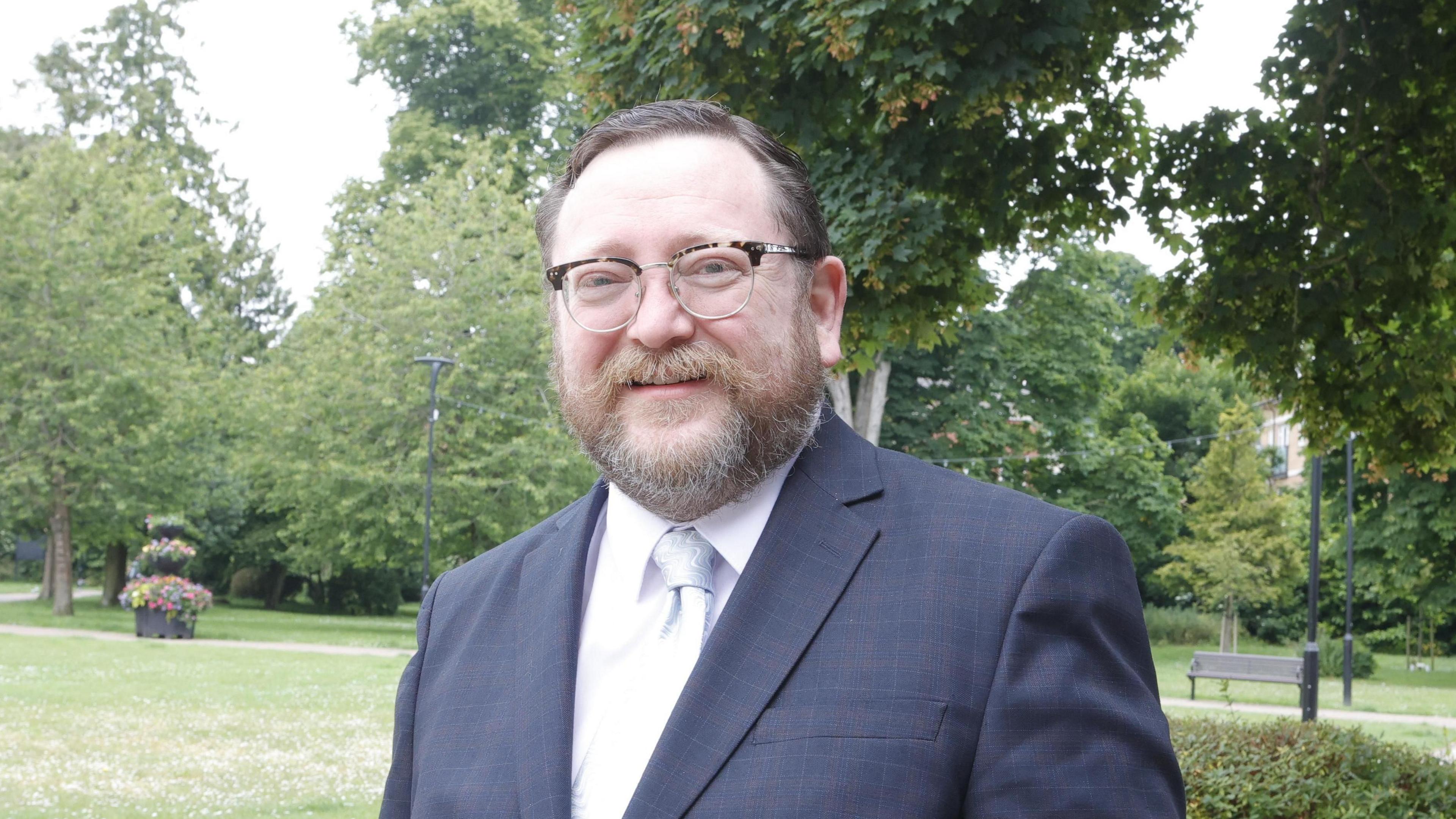 A man with brown hair, beard and glasses looking into the camera as he stands in a park with trees and grass in the background. He is wearing a blue suit with a white shirt and a tie.