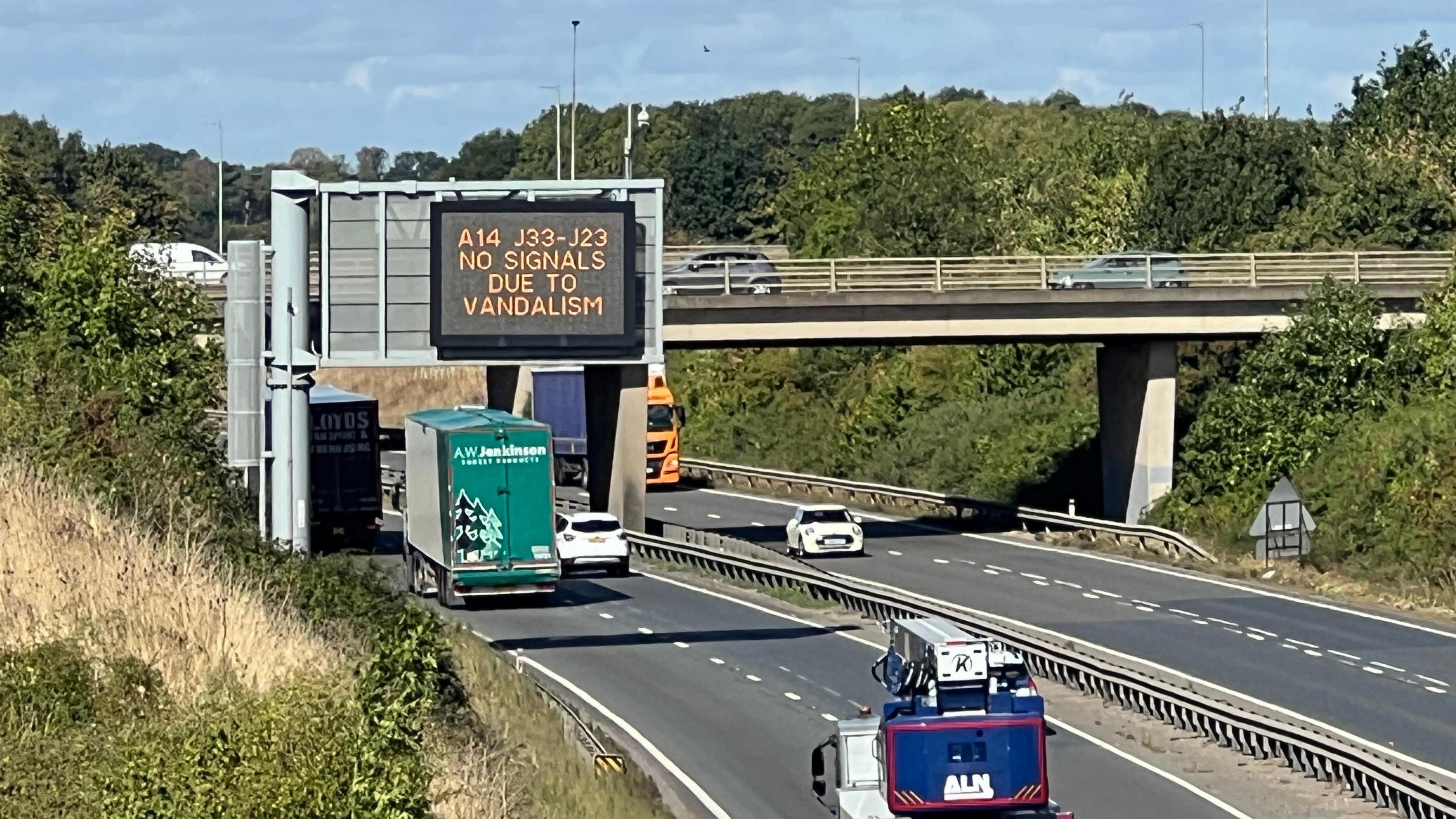 Traffic is travelling on the A14 under a large electronic sign. In yellow writing it says 'A14 J33-j23, No signals due to vandalism'. The image has been taken looking down onto the A14 from a footbridge.
