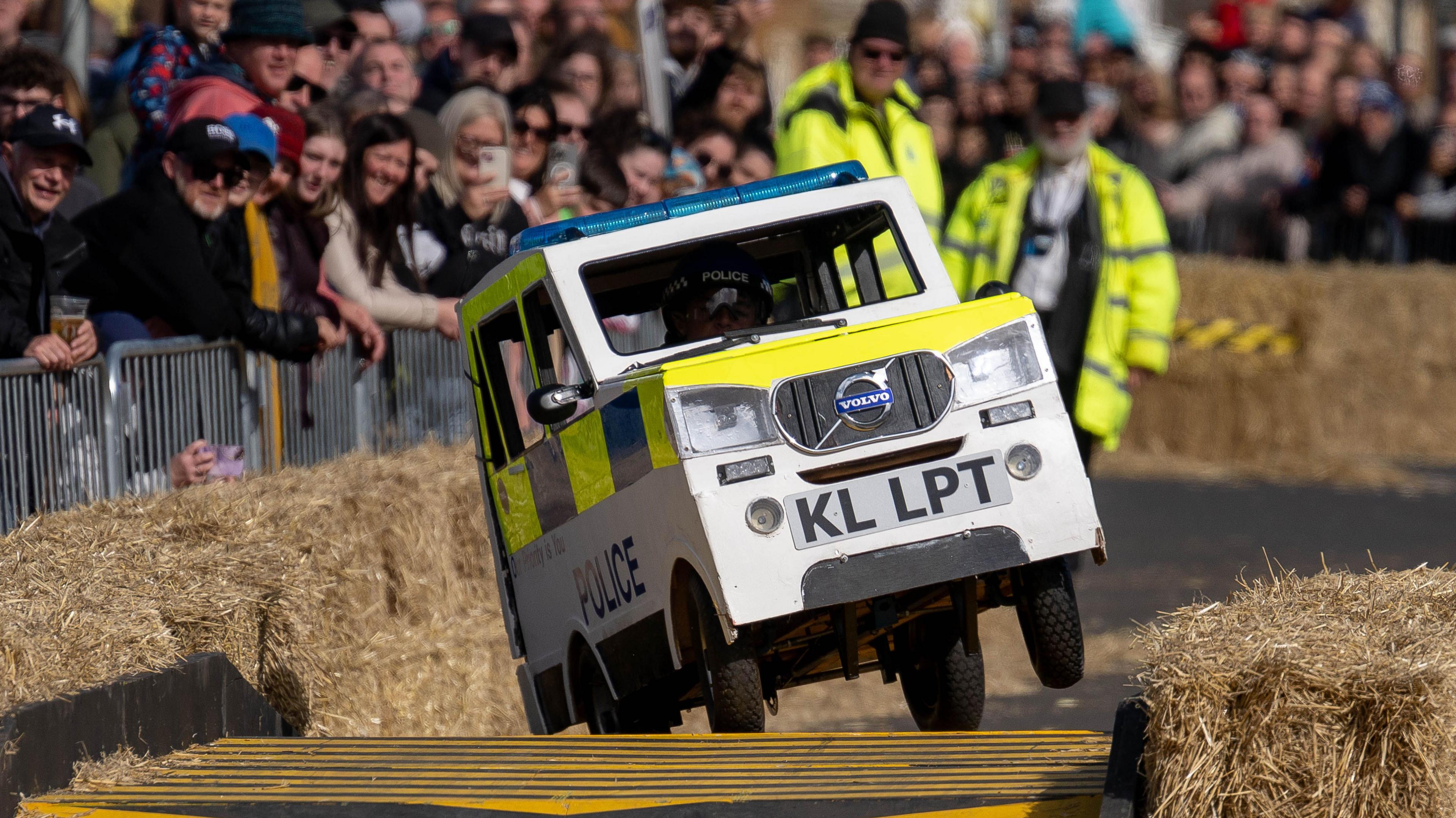 A hand-built kart which has been designed to look like a white police van with painted yellow and blue fluorescents rectangles on the side and blue sirens on top.
