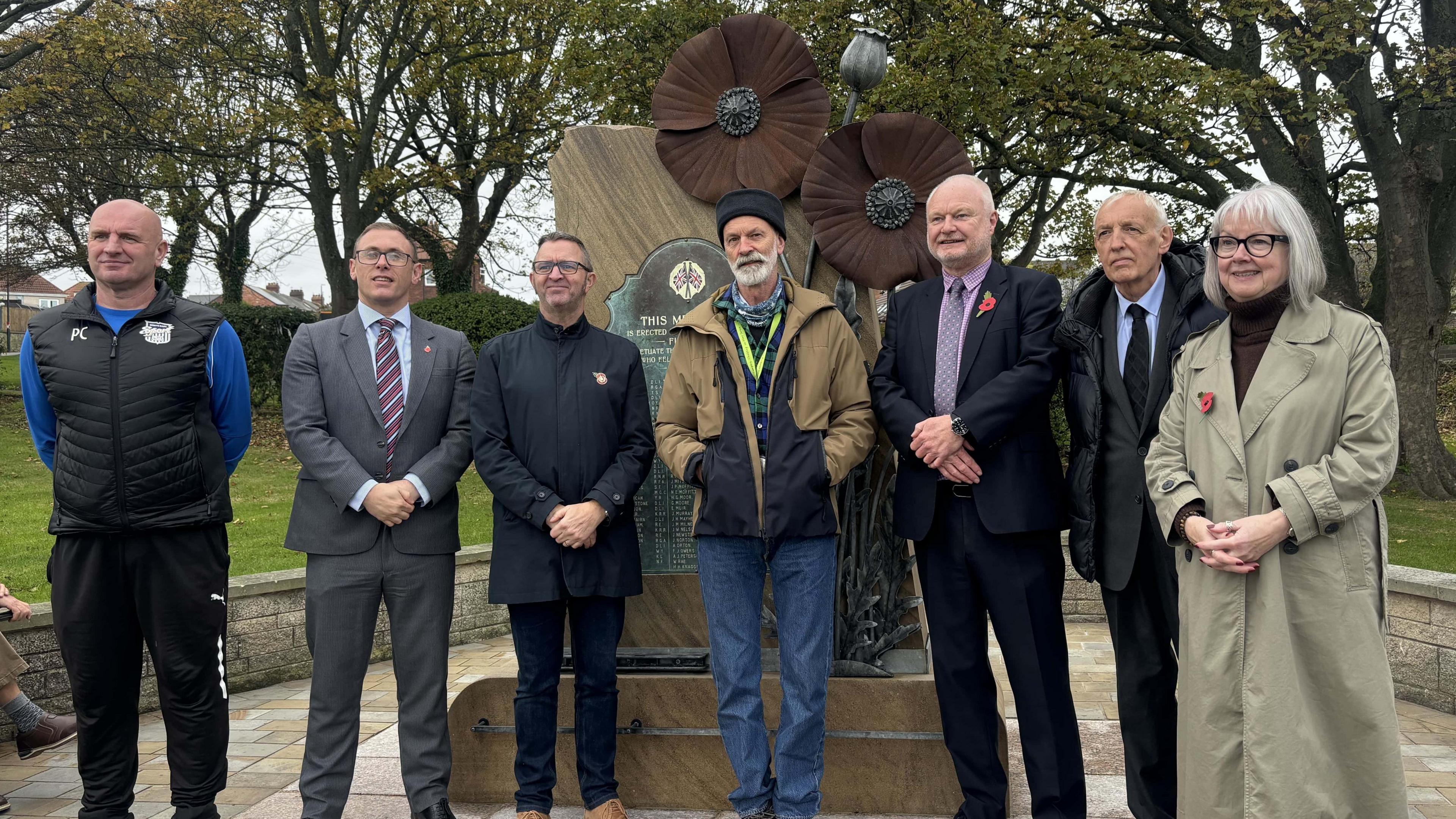 A group of six men and one woman standing in front of a sculpture which includes two large poppies in dark brownish red. Colin Wilbourn is in the middle and Michael Hartnack is next to him on the right. Mr Wilbourn has a white beard and a black hat. He is wearing a brown jacket. Hartnack is wearing a suit with a red poppy. He has a tie on.